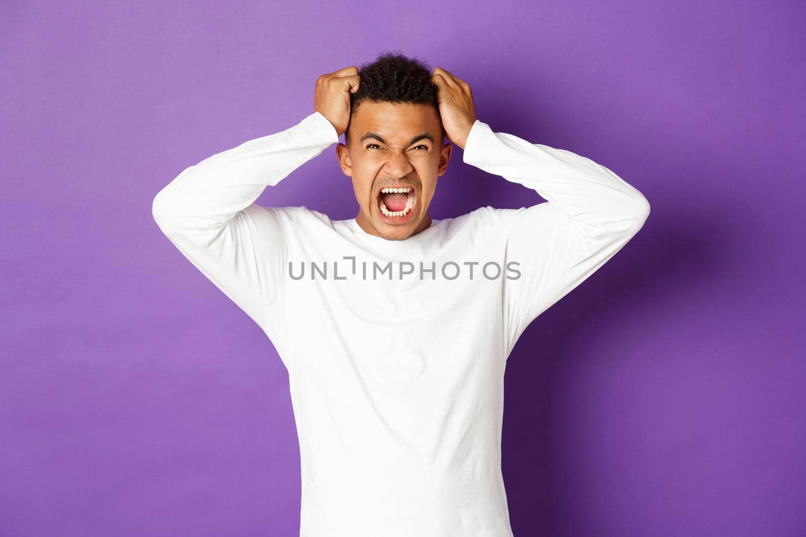 Portrait of tensed and distressed african-american guy losing temper, shouting and ripping hair on head, looking angry up, standing over purple background.