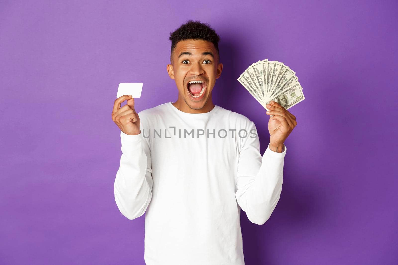 Image of handsome african american guy, looking pleased and smiling, showing credit card and money, standing over purple background.