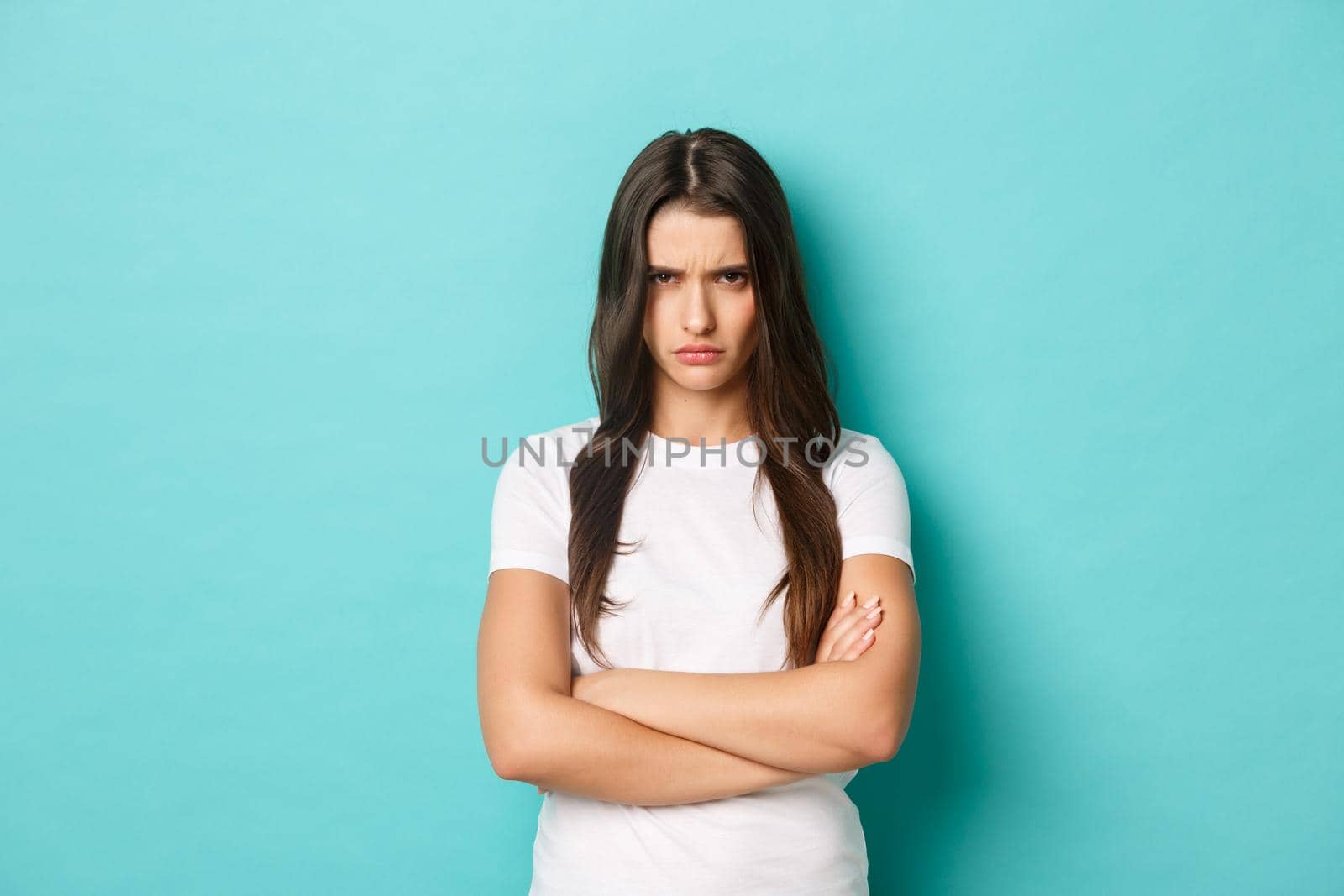 Image of angry young woman in white t-shirt, mad at you, frowning and looking from under forehead, cross arms on chest and sulking, standing over blue background.