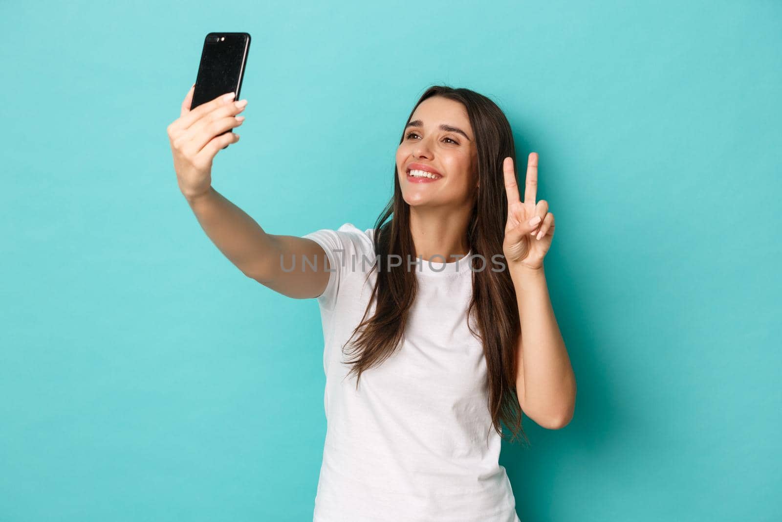 Portrait of beautiful happy woman in white t-shirt, taking selfie with peace sign, smiling at mobile phone, standing over blue background.
