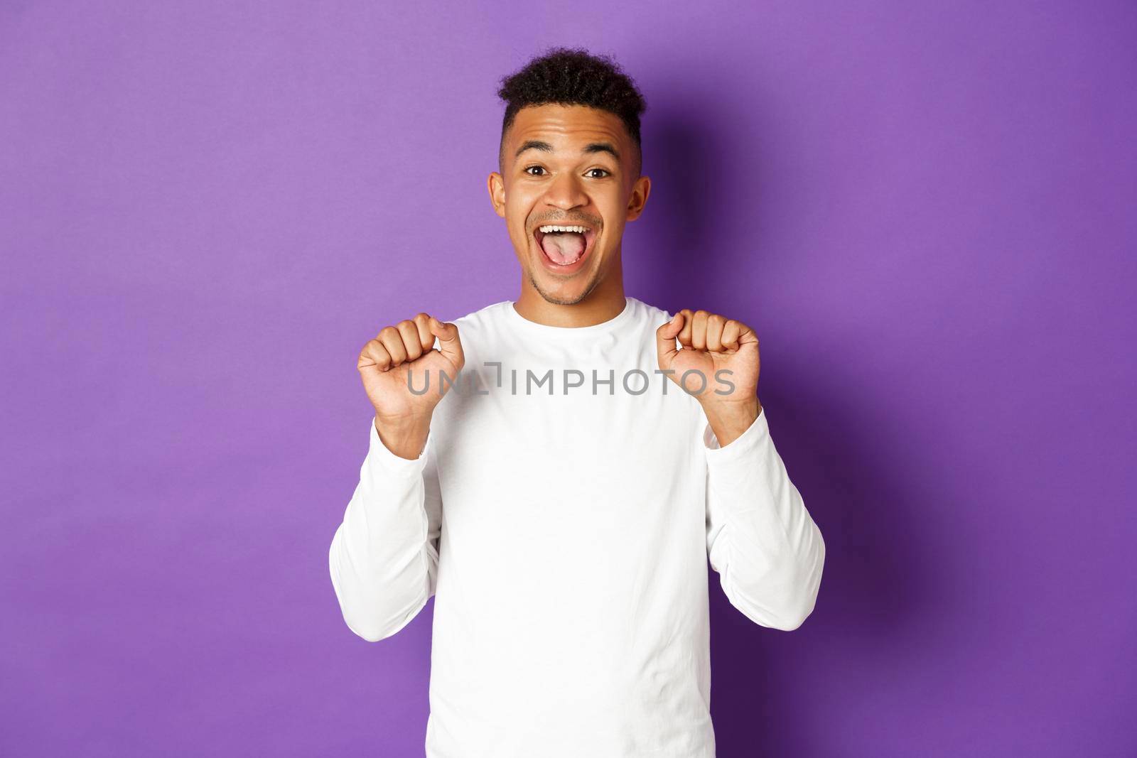 Image of happy and cheerful african-american man in white sweatshirt, jumping from joy, celebrating success or achievement, standing against purple background by Benzoix