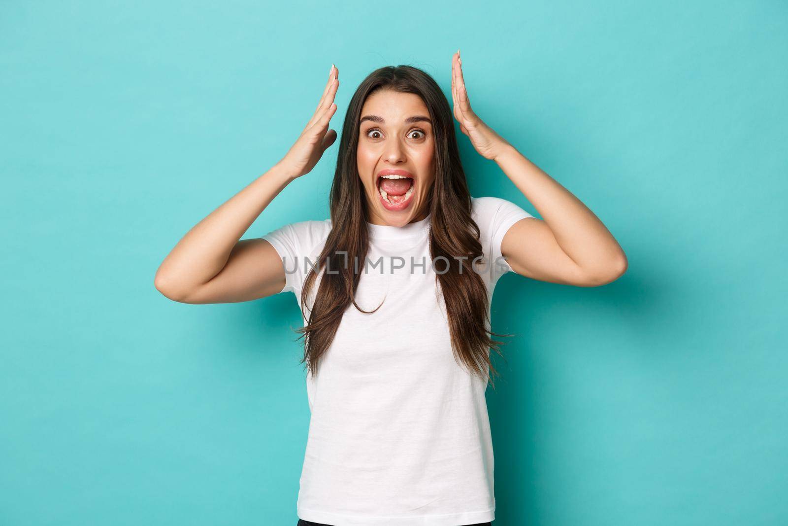 Image of scared and alarmed brunette girl in white t-shirt, shouting and holding hands over head, standing in panic over blue background.