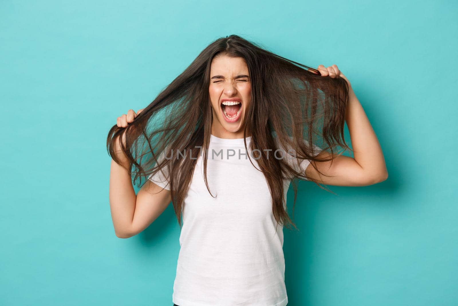 Image of distressed and angry brunette woman in white t-shirt, ripping hair off head and screaming, standing frustrated over blue background.