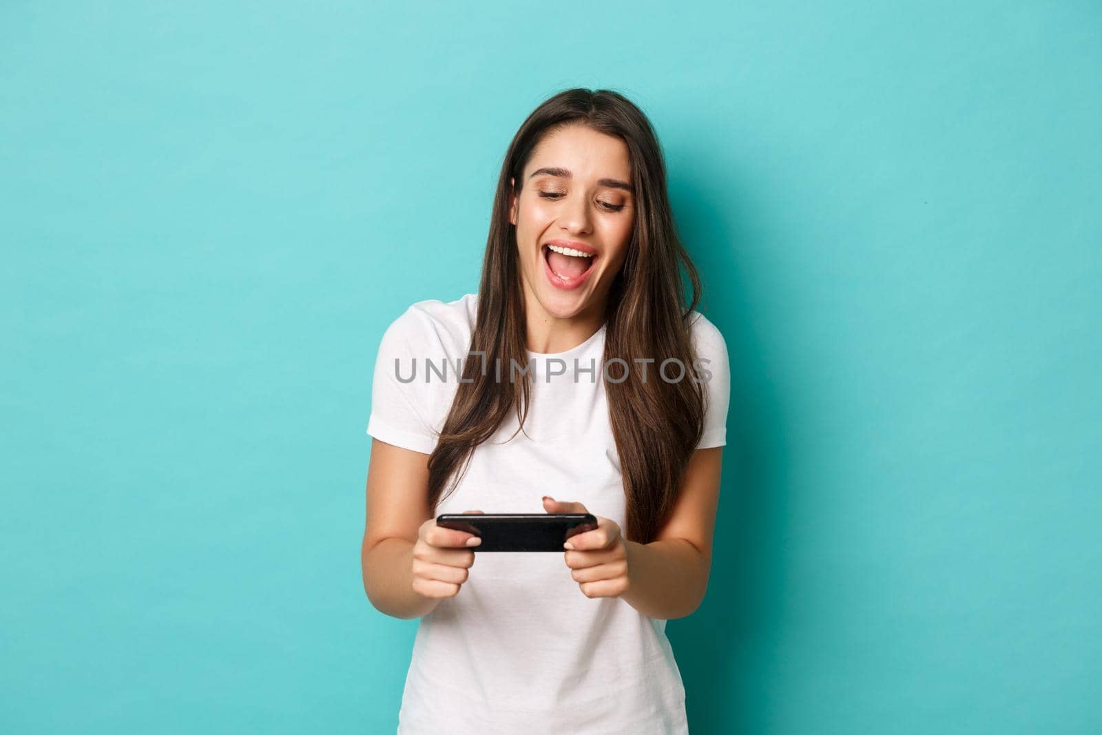 Image of happy young woman in white t-shirt, playing mobile phone game and smiling, standing over blue background.