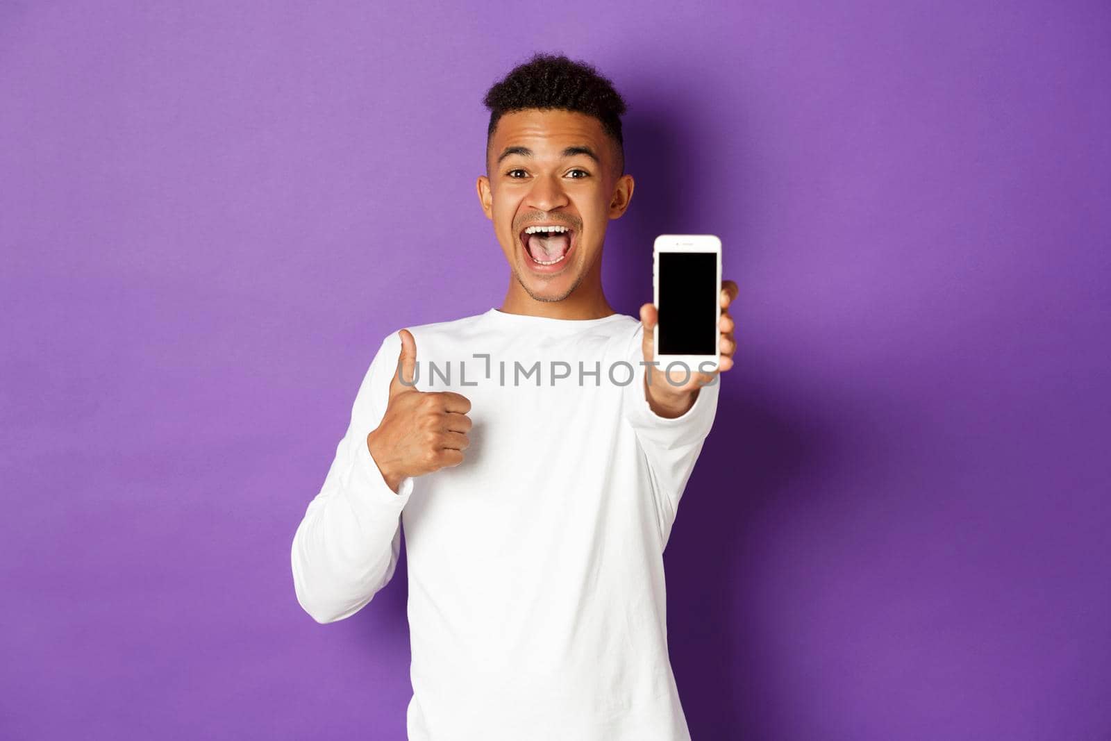 Image of african-american guy in white sweatshirt, smiling and showing thumbs-up in approval, showing mobile phone screen, standing over purple background by Benzoix