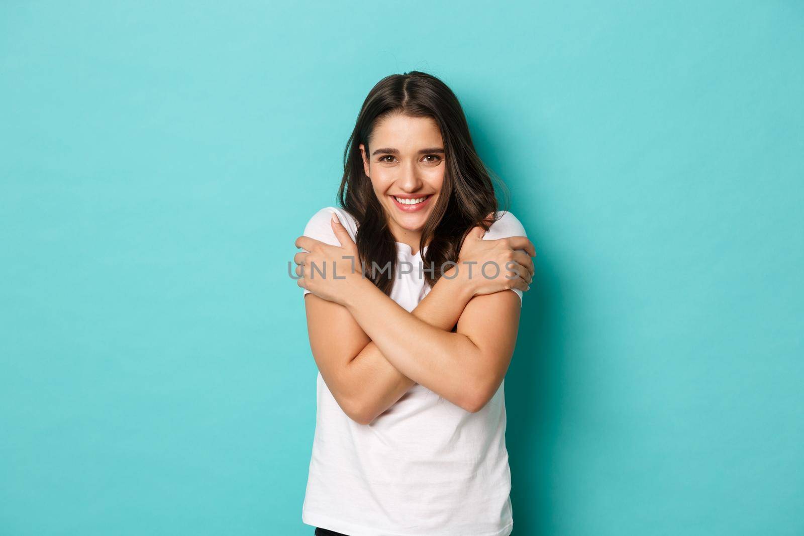 Image of cute smiling girl in white t-shirt, embracing herself, feeling chilly, standing over blue background.