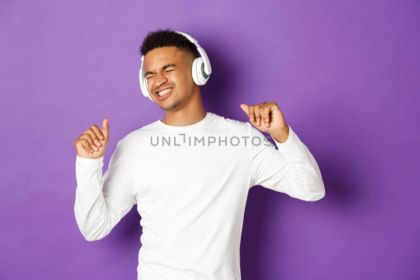 Portrait of handsome modern african-american guy, listening music in headphones and dancing carefree, standing over purple background by Benzoix