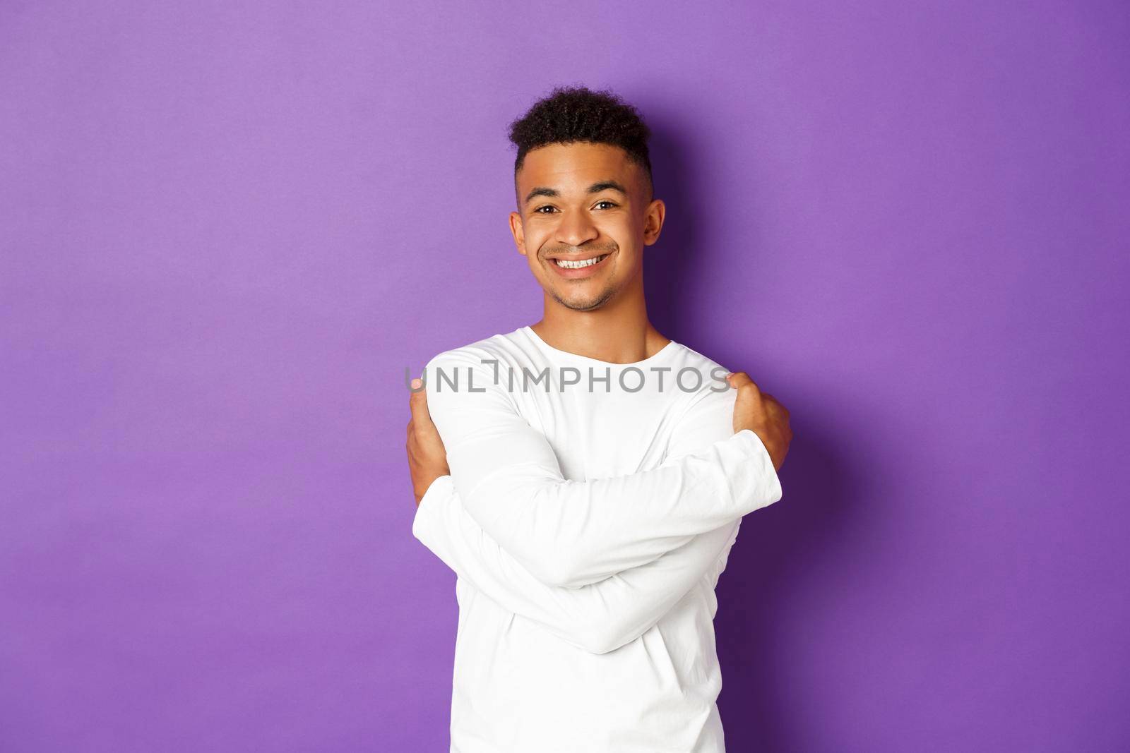 Cheerful african-american man in white sweatshirt, embracing himself and smiling, standing happy over purple background by Benzoix