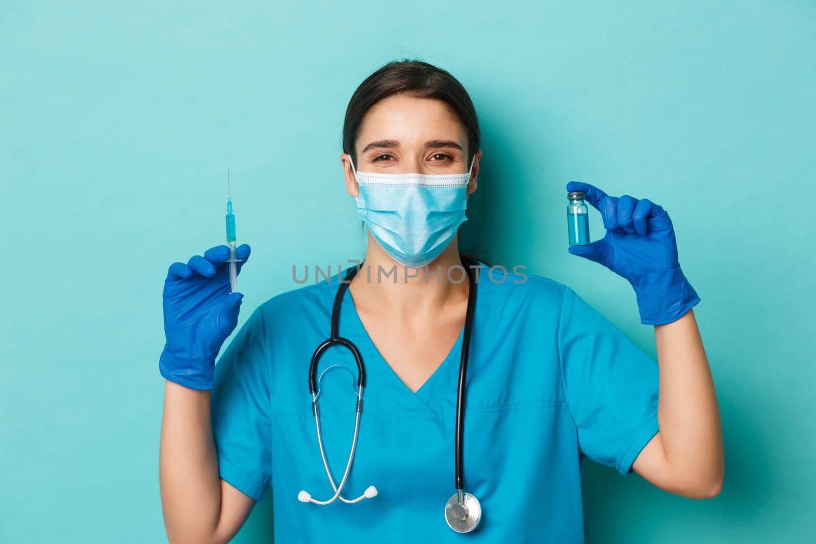 Concept of covid-19 and quarantine concept. Cheerful smiling female doctor in medical mask, latex gloves and scrubs, showing syringe filled with coronavirus vaccine, standing over blue background by Benzoix