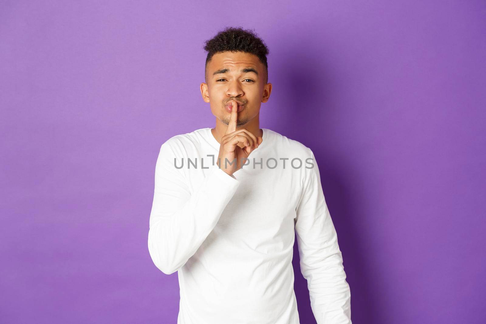 Young worried african-american guy asking to keep quiet, making hush sign and looking at camera, need silence, standing over purple background.