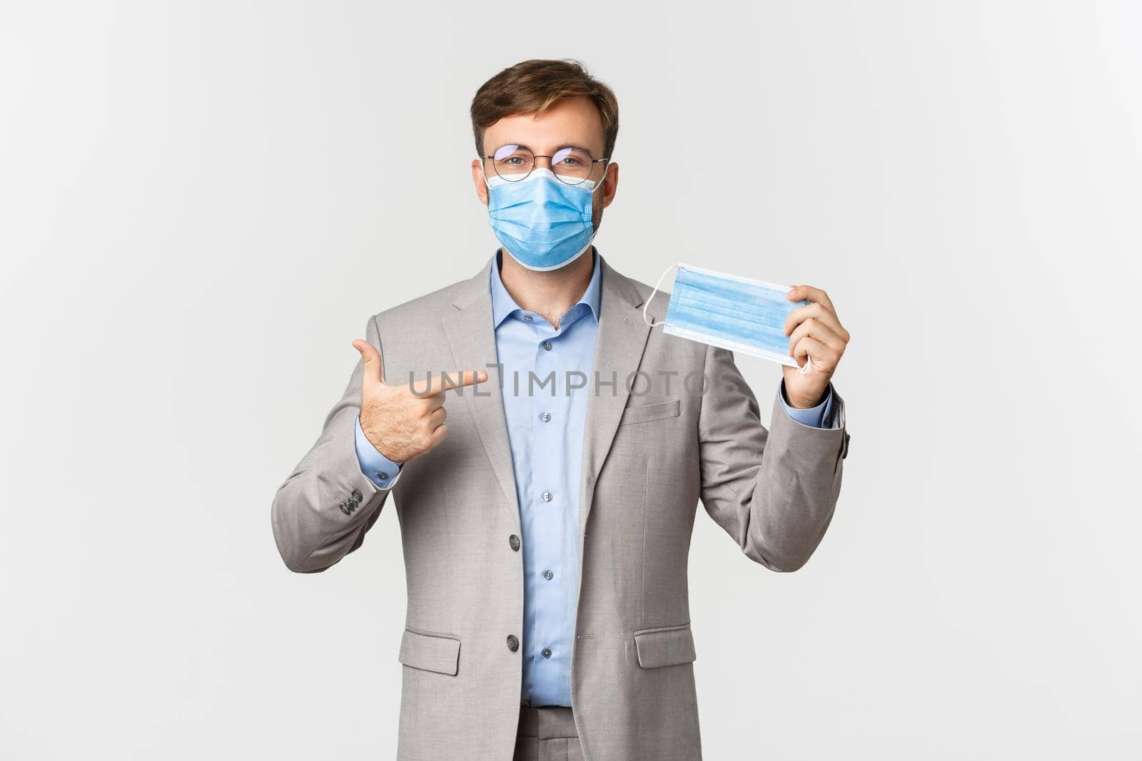 Concept of work, covid-19 and social distancing. Image of male employer in gray suit, encourage using medical mask in office, standing over white background.