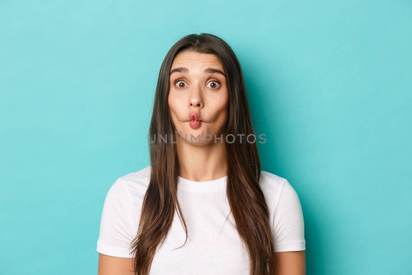 Close-up of silly brunette woman, sucking lips and showing funny faces, standing over blue background.