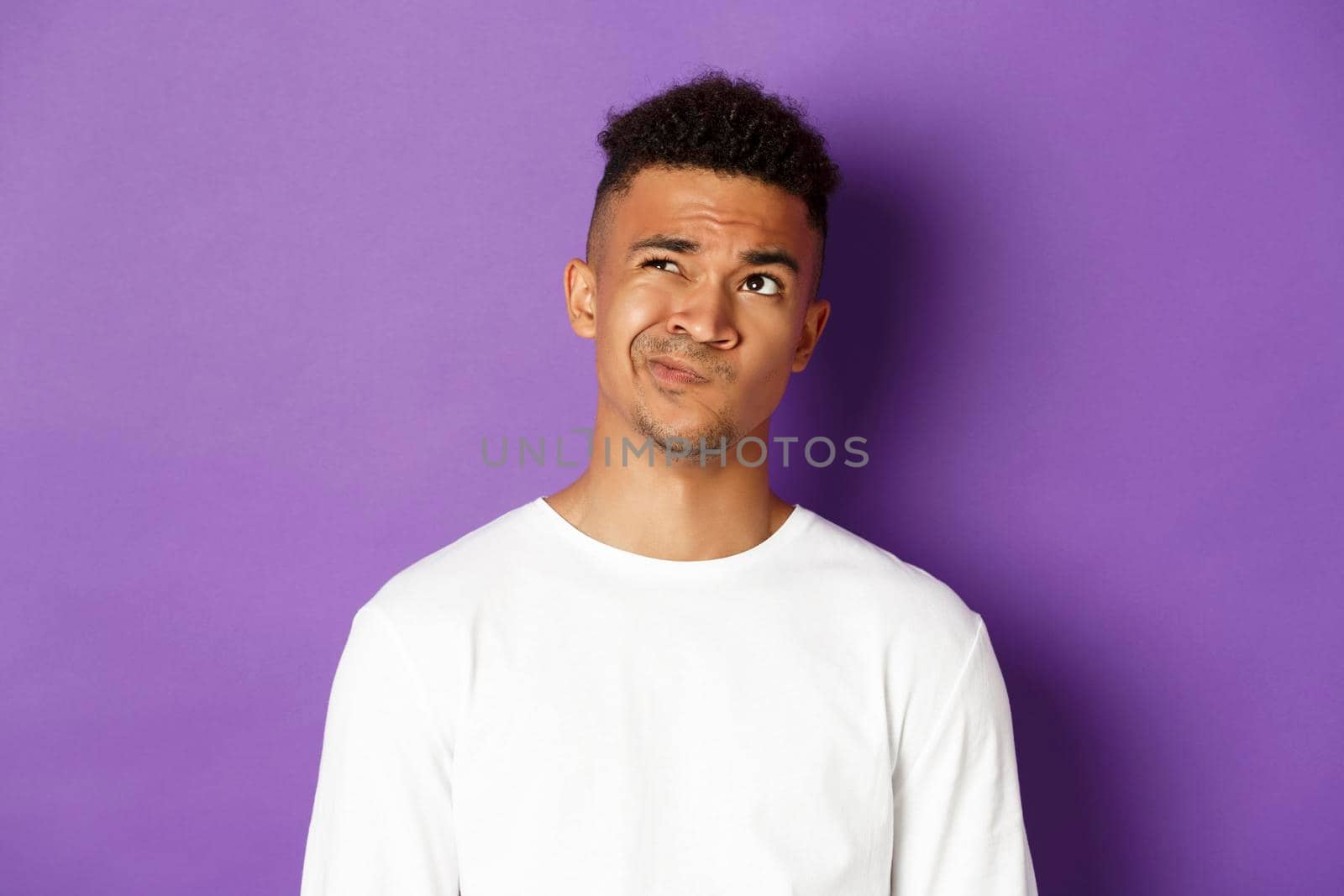 Close-up of indecisive african-american man making choice, looking perplexed at upper left corner, thinking about something complicated, standing over purple background.