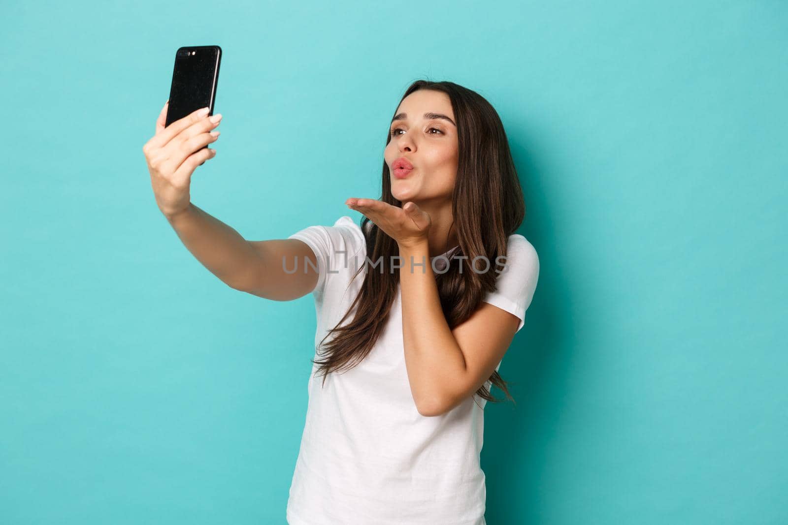 Portrait of attractive modern woman in white t-shirt, sending air kiss and mobile phone camera, taking selfie against blue background by Benzoix
