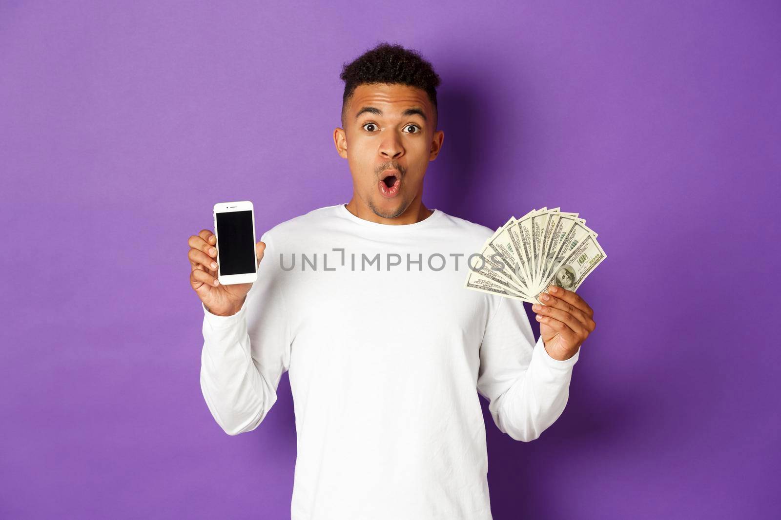 Portrait of handsome african-american man in white sweatshirt, showing money and mobile phone screen, looking amazed, standing over purple background.