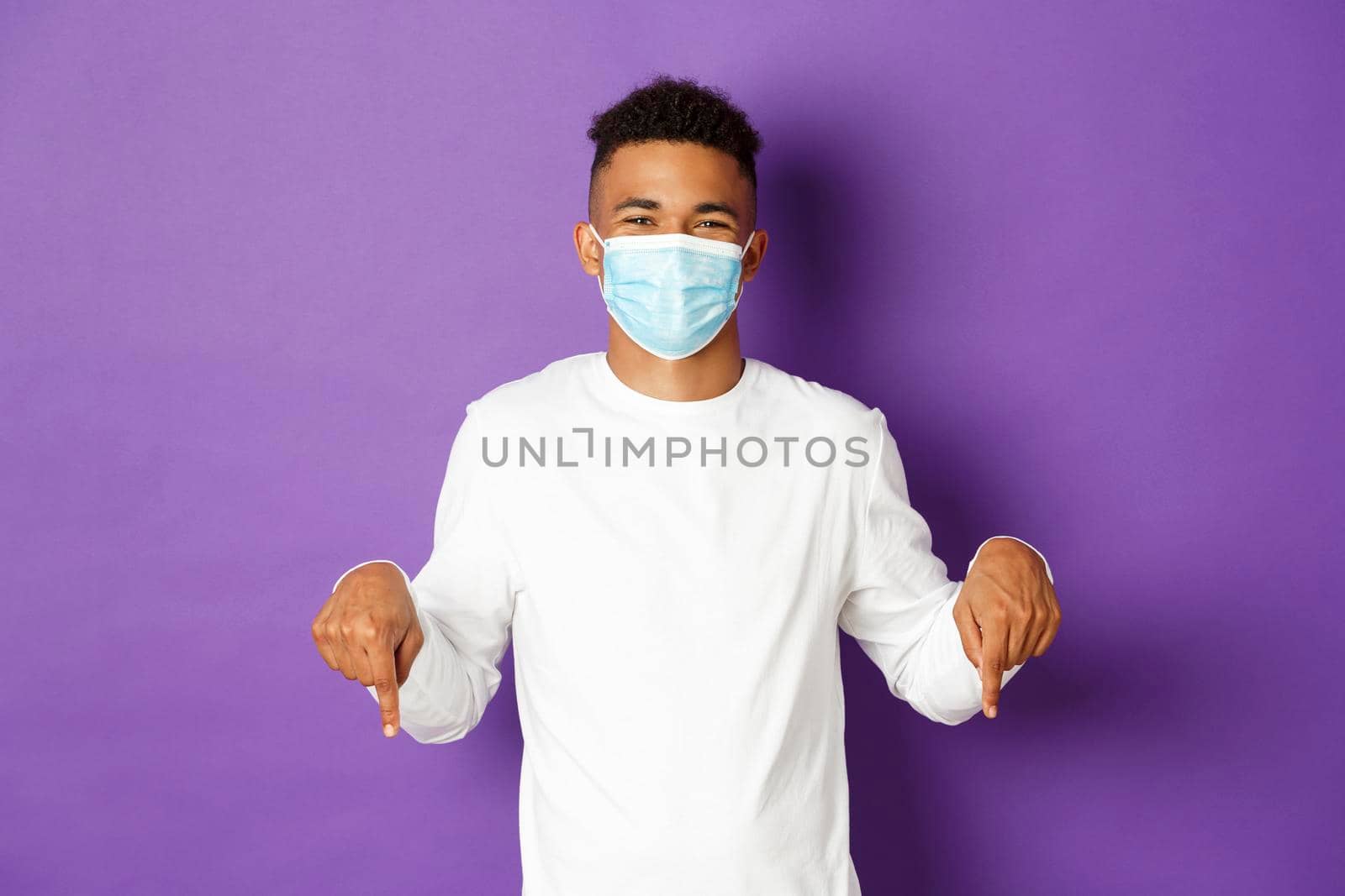 Concept of coronavirus, quarantine and lifestyle. Handsome african-american guy in medical mask showing promo, pointing fingers down at logo and smiling, standing over purple background by Benzoix