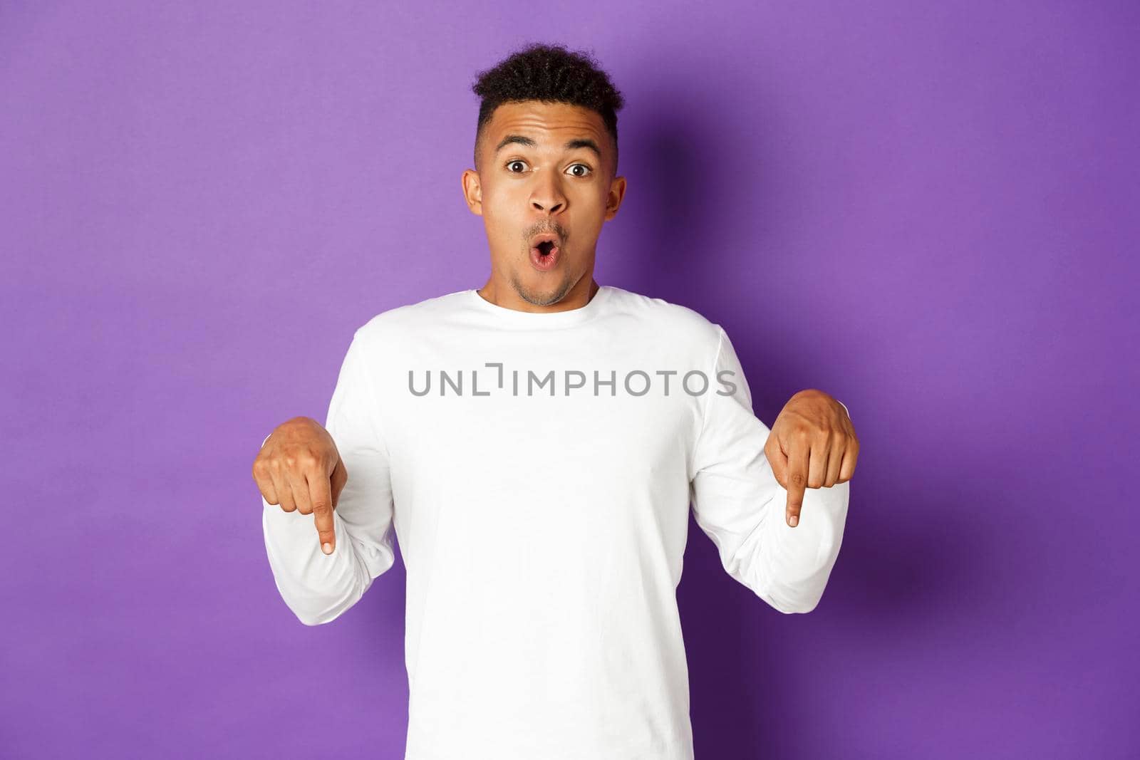 Image of handsome african-american man looking surprised, saying wow and pointing fingers down at something amazing, showing promo, standing over purple background by Benzoix