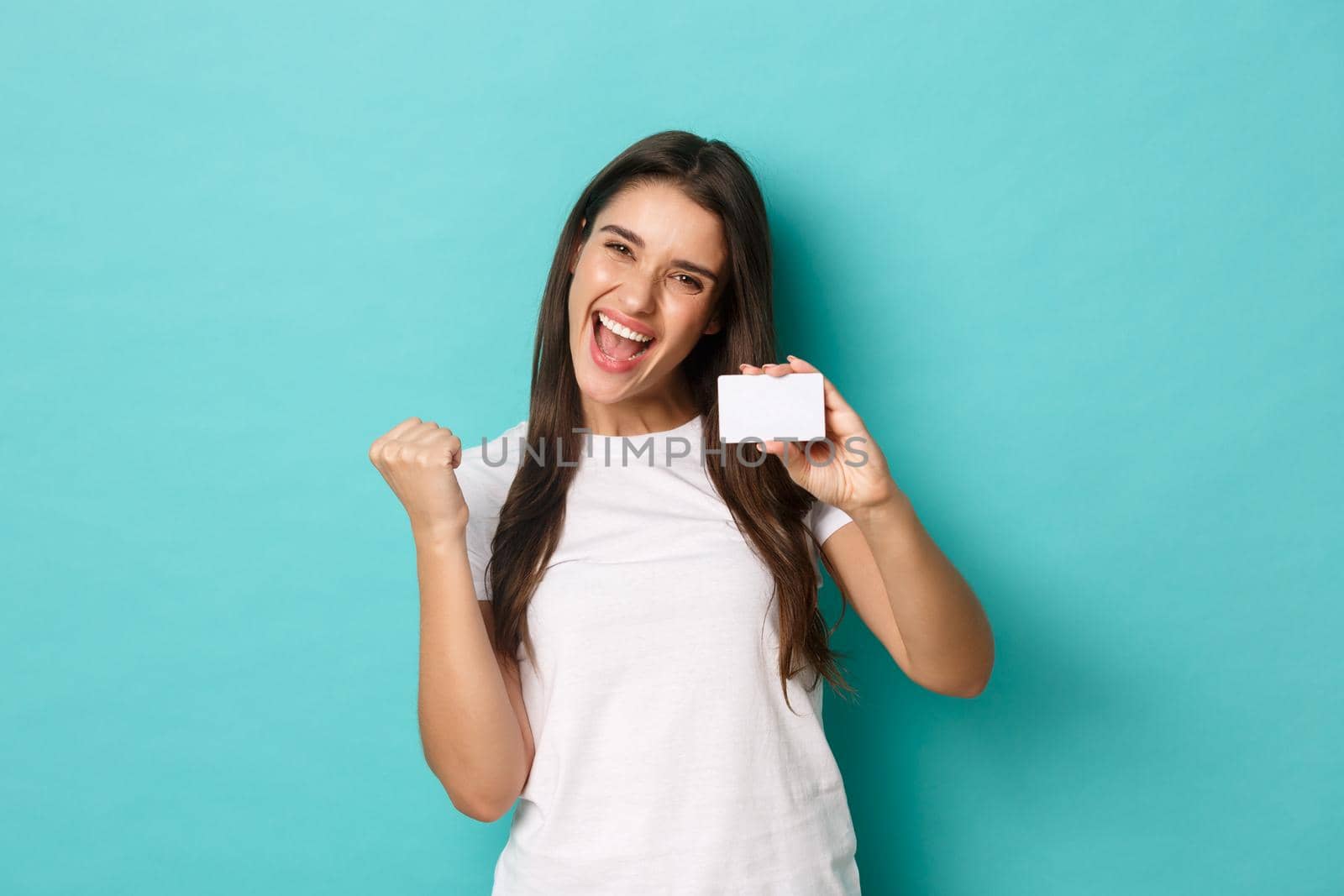 Image of satisfied ecstatic woman, wearing white t-shirt, rejoicing and showing credit card, standing pleased over blue background by Benzoix