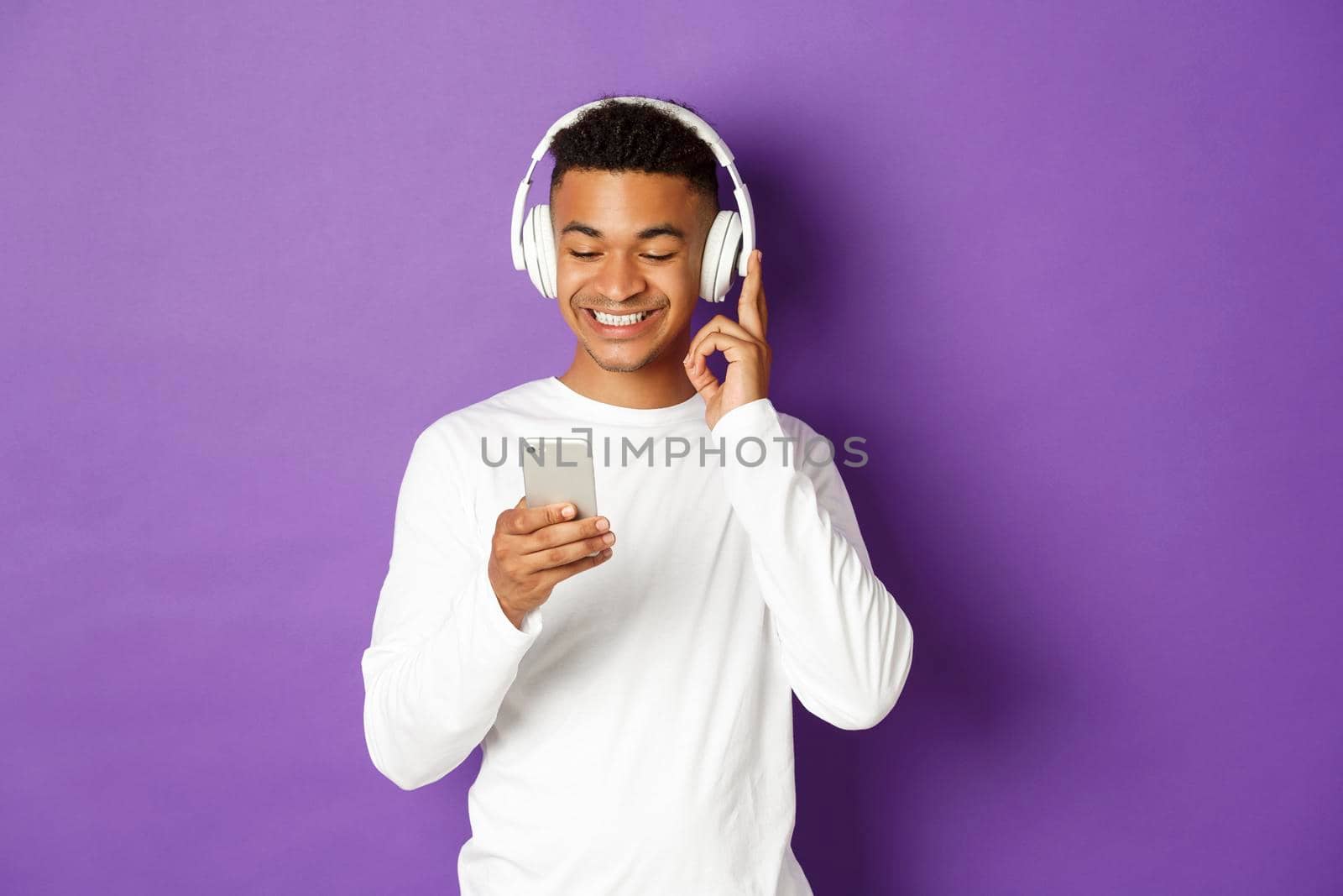 Image of modern african-american guy in white sweatshirt, listening music in headphones and looking at mobile phone, standing over purple background.