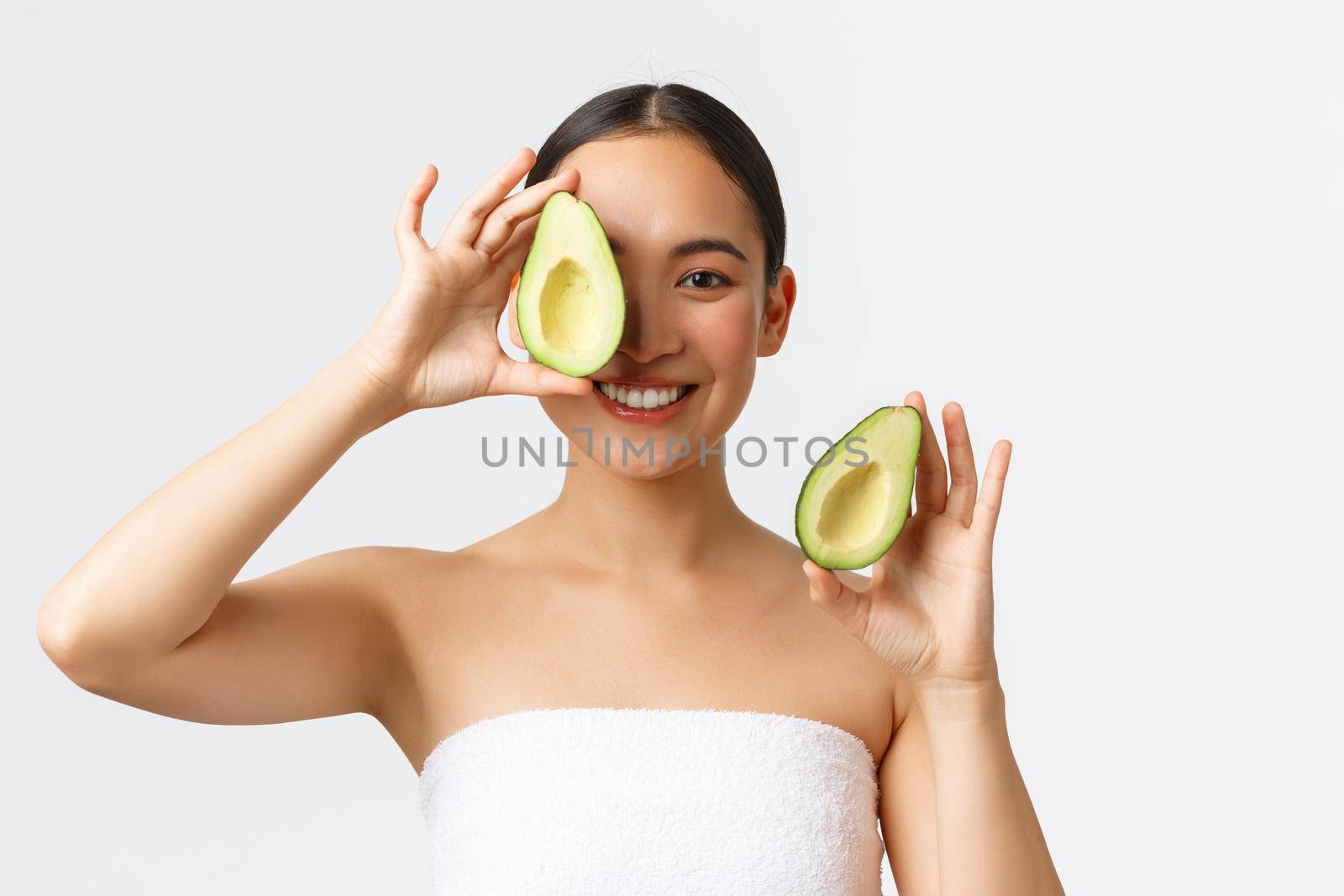 Beauty, personal care, spa and skincare concept. Close-up of tender feminine asian woman in bath towel, smiling and showing avocado near face, advertisement of face mask, cleanser or cream.