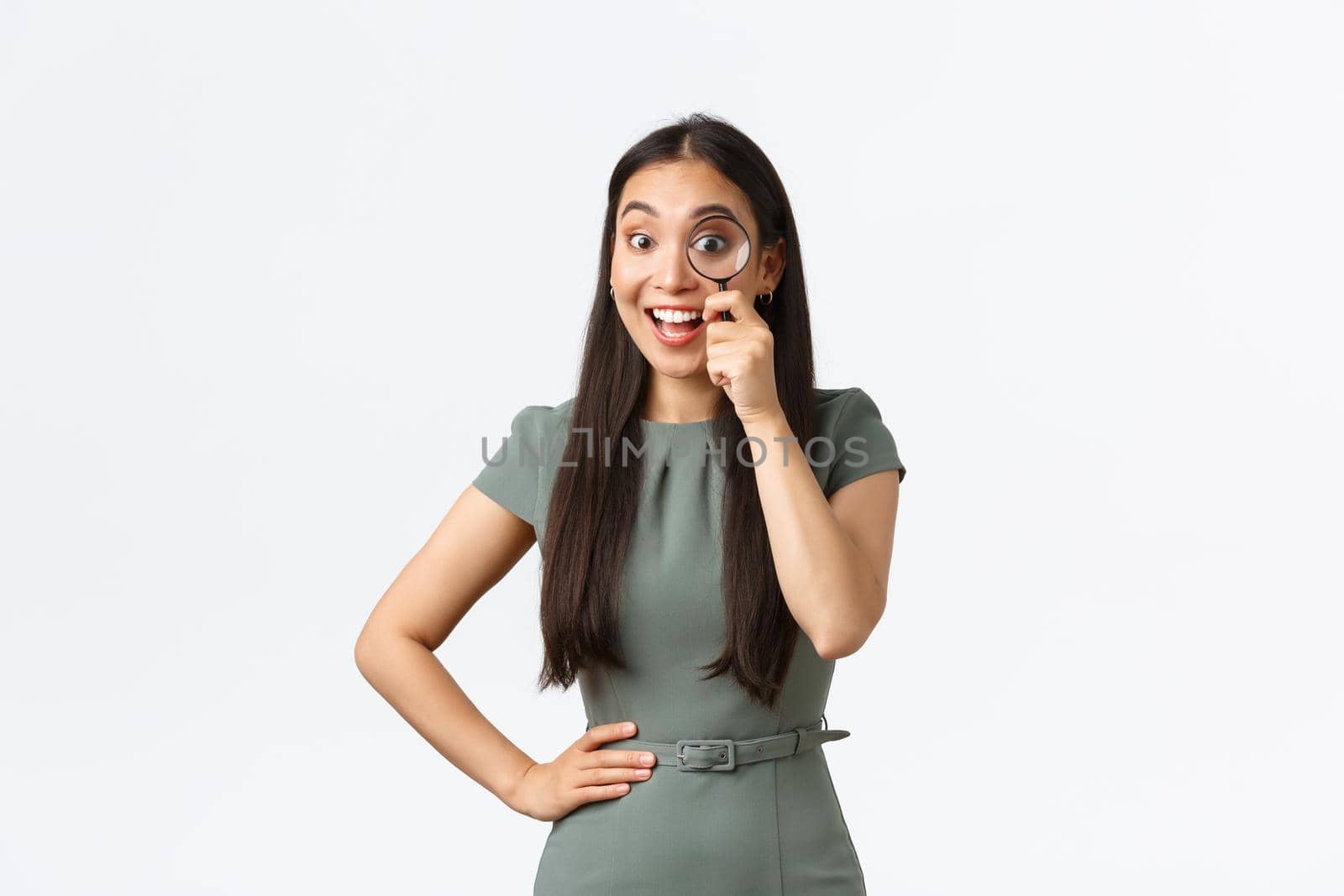 Small business owners, women entrepreneurs concept. Excited happy asian woman found something good, looking through magnifying glass with pleased smile, searching for product, white background.