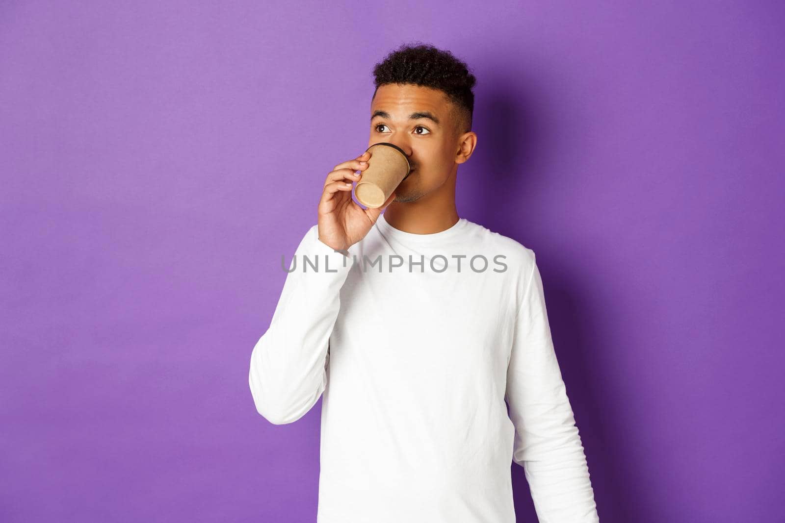 Portrait of young modern african-american guy drinking coffee, looking at upper left corner, standing over purple background by Benzoix