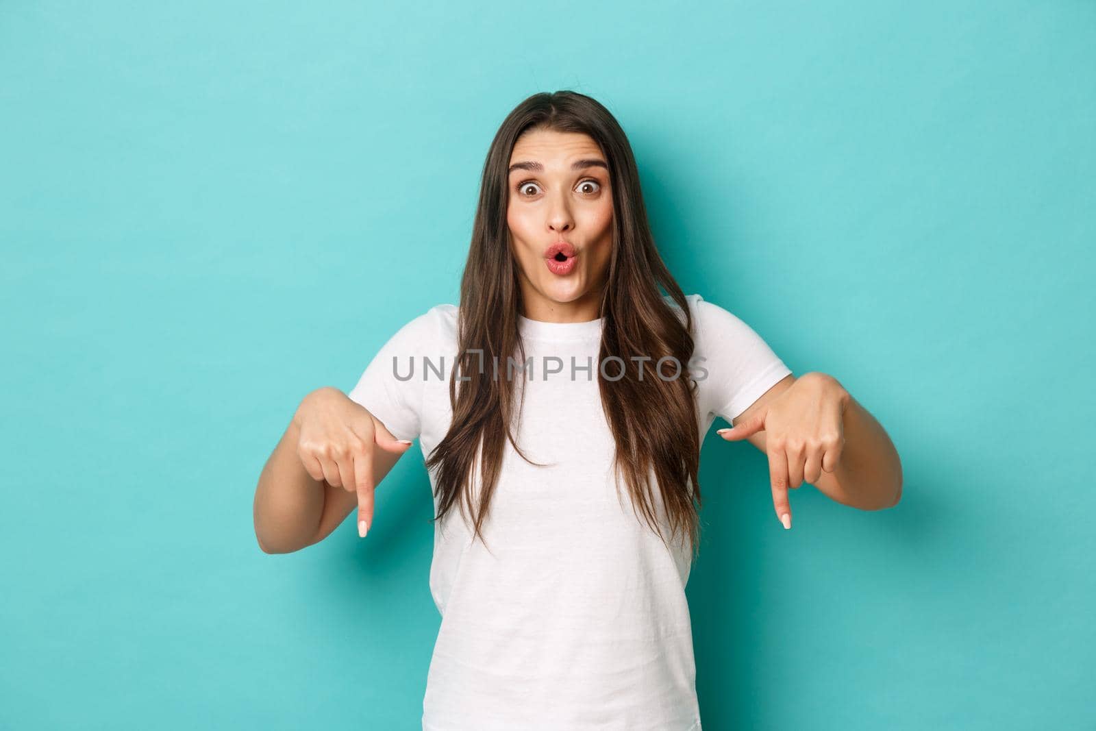 Image of excited beautiful woman showing something awesome, pointing fingers down and looking amazed, standing over blue background.