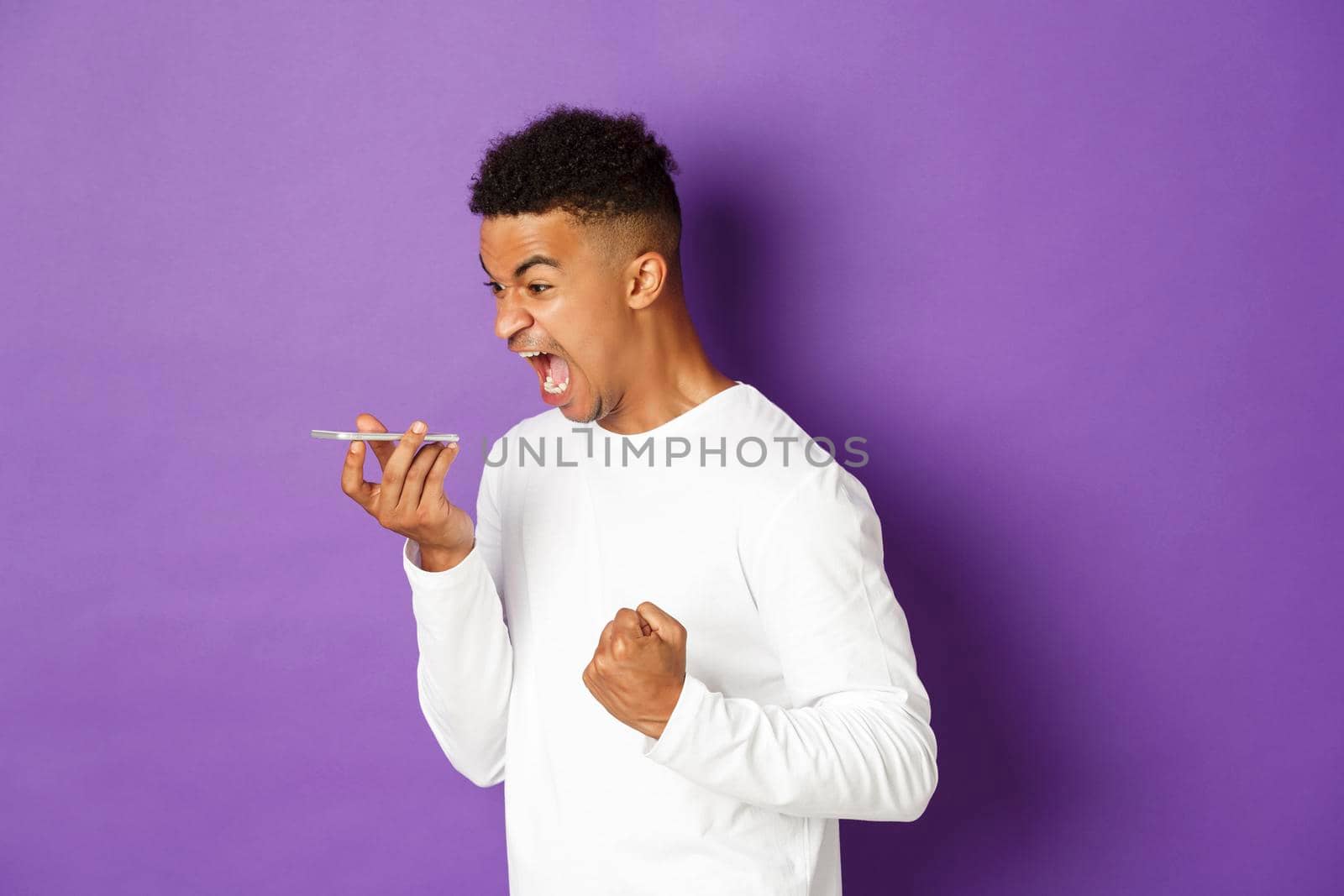 Image of angry african-american man shouting at speakerphone, looking angry and pissed-off, standing with mobile phone over purple background.