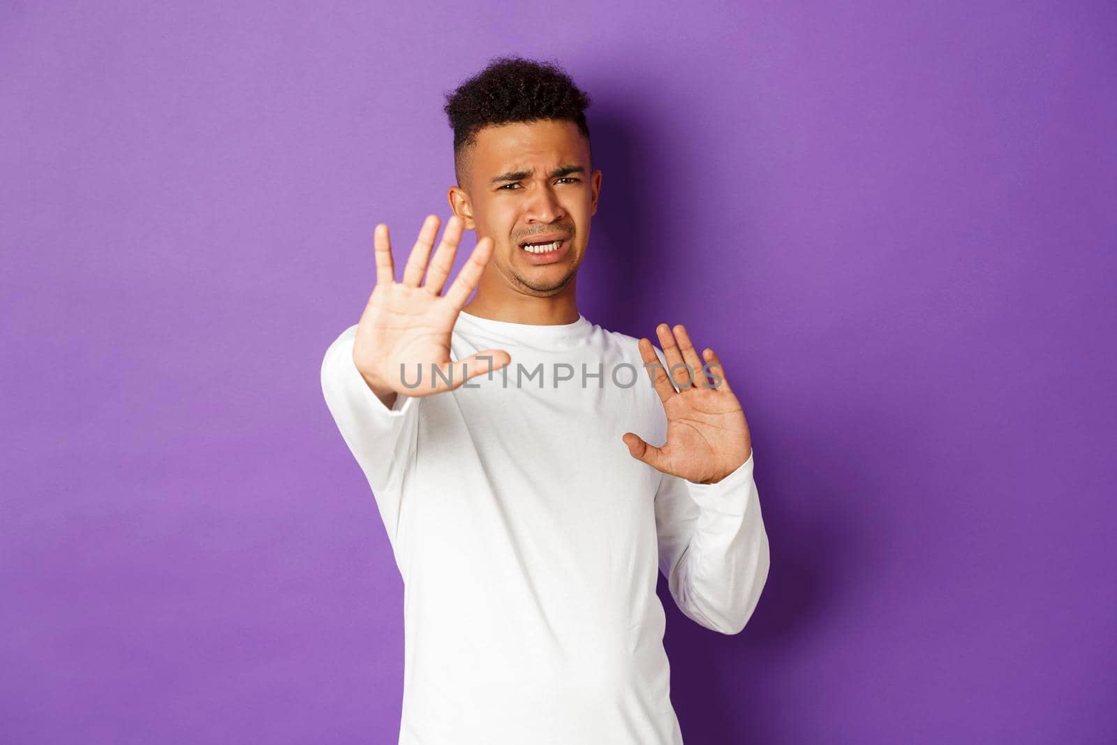 Image of african-american young man feeling uncomfortable, asking to stop, defending himself with raised arms and frowning, standing against purple background.