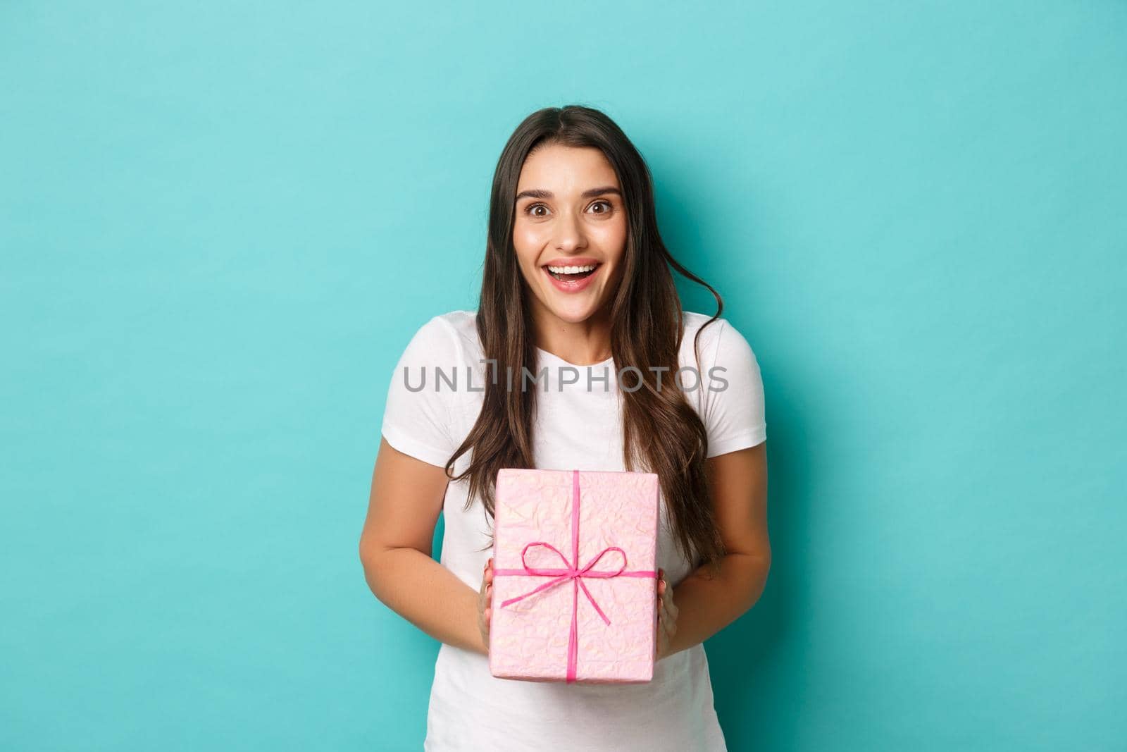 Concept of holidays and celebration. Image of excited woman looking happy, smiling and receiving gift wrapped in pink box, standing over blue background.