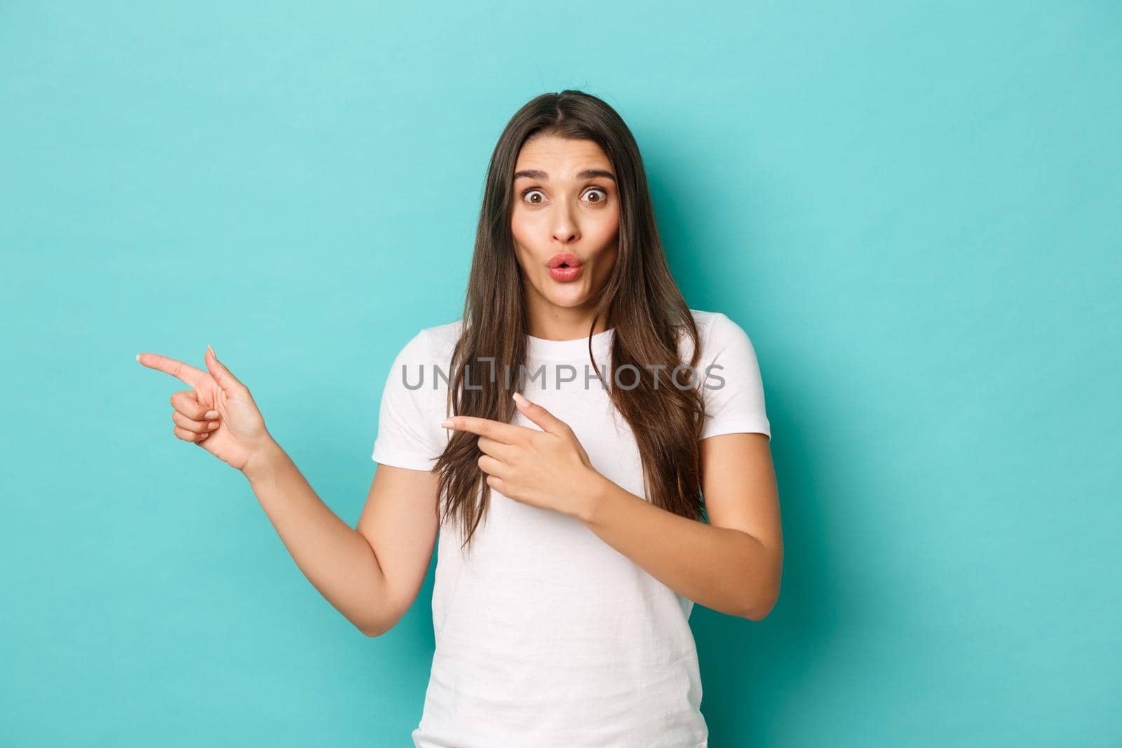 Portrait of beautiful surprised woman in white t-shirt, pointing fingers left at copy space, showing logo, standing over blue background.