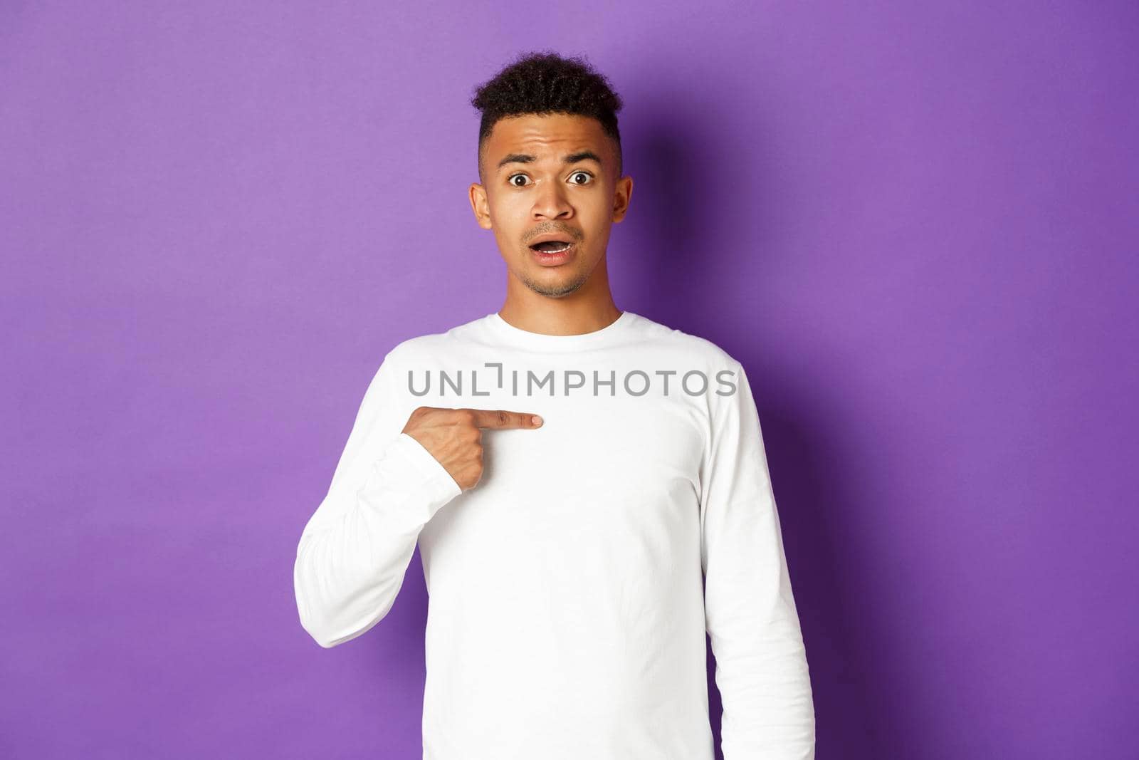 Portrait of confused and nervous african-american young man, pointing at himself, being chosen, standing over purple background.