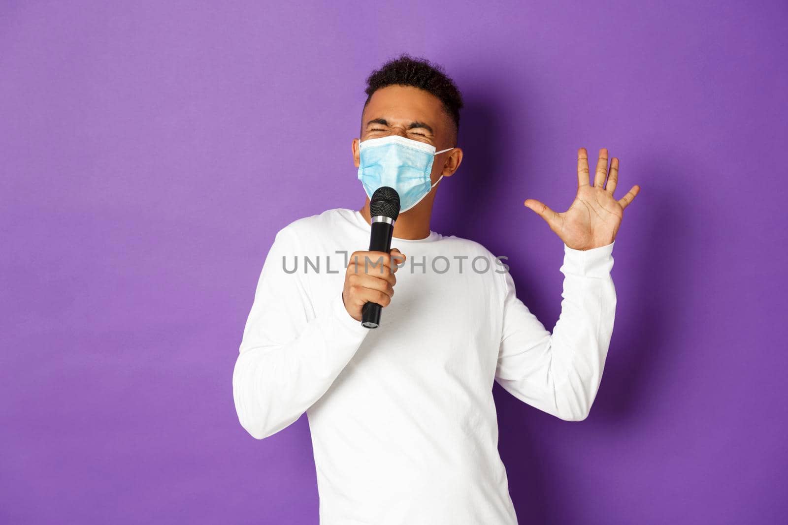 Concept of covid-19, pandemic and social distancing. Cheerful african-american guy having fun at karaoke, wearing medical mask and singing in microphone, standing over purple background.