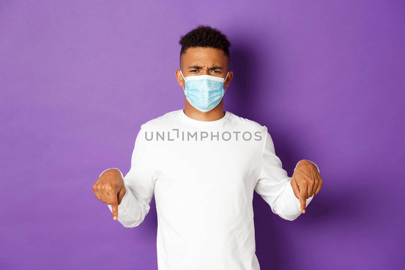 Concept of coronavirus, quarantine and lifestyle. Frustrated and angry african-american man in medical mask, complaining at logo, pointing fingers down, standing over purple background.