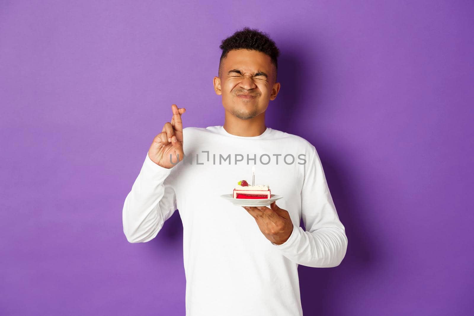 Image of excited african-american man celebrating birthday, making wish, holding b-day cake and crossing fingers, standing over purple background by Benzoix