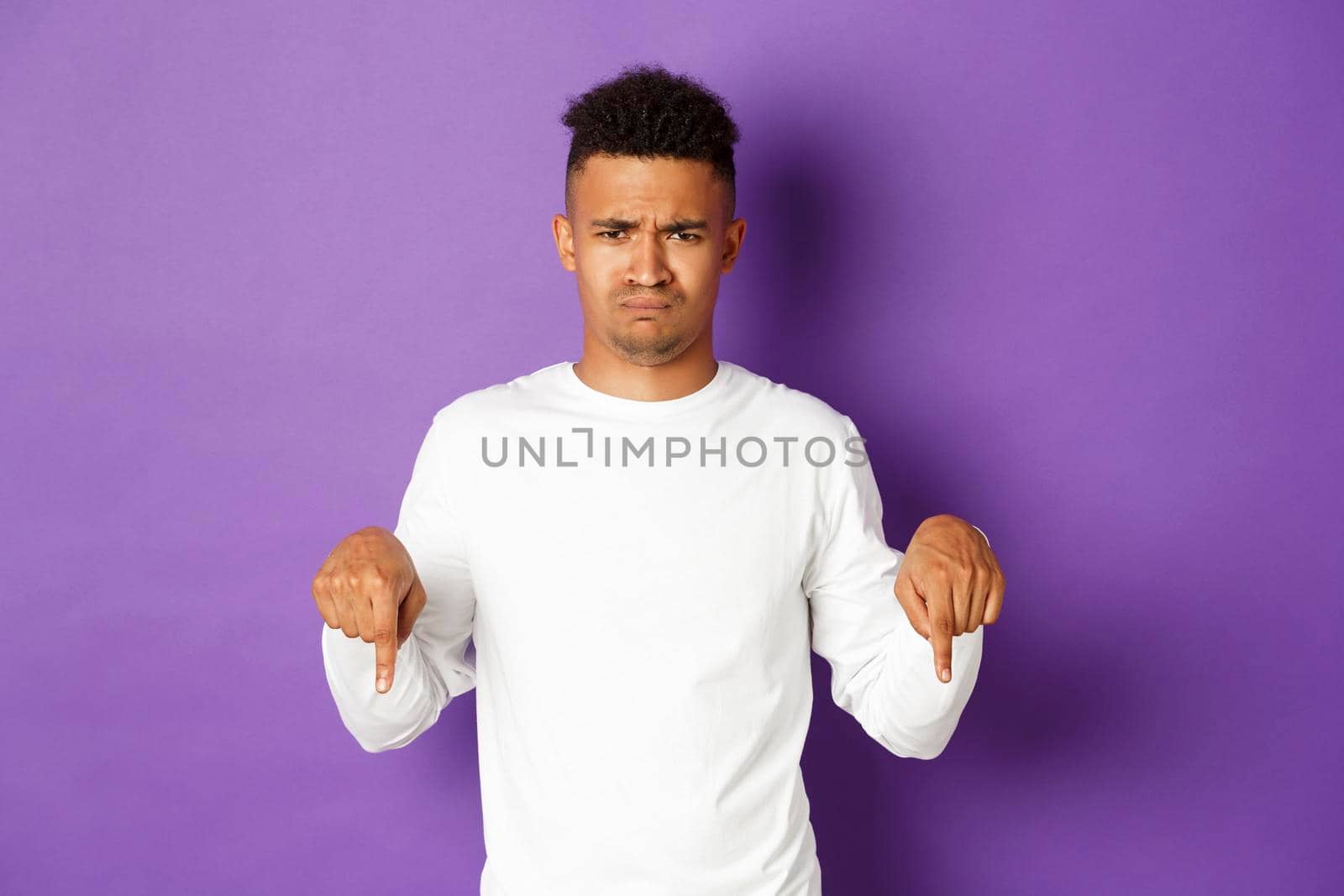 Portrait of silly upset african-american guy, frowning and sulking, complaining, pointing fingers down at something disappointed, standing over purple background.