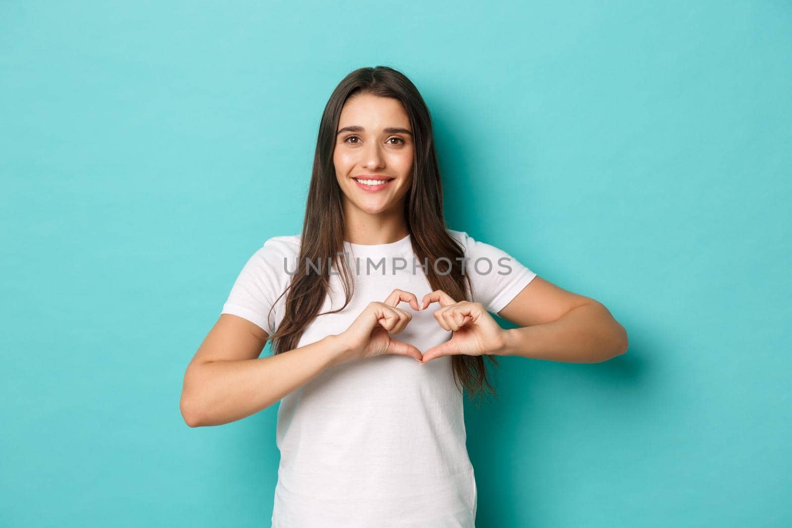Image of lovely brunette girl in white t-shirt, smiling happy and showing heart sign, express love and sympathy, standing over blue background.