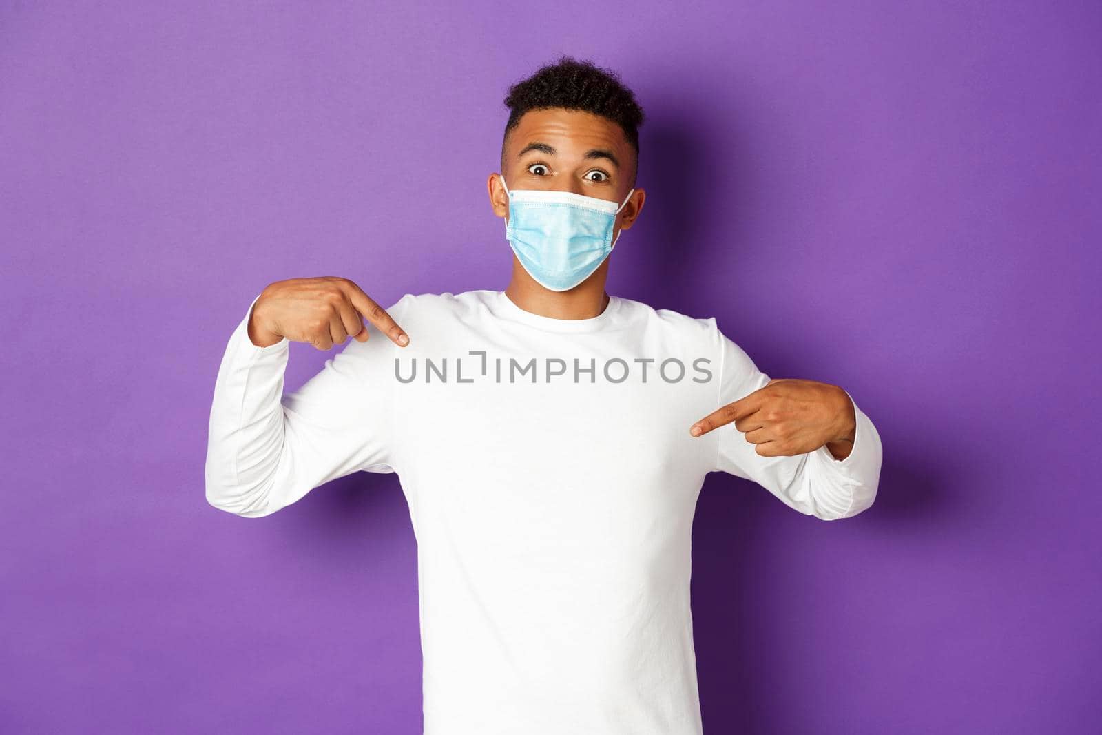 Concept of coronavirus, quarantine and lifestyle. Excited african-american guy in medical mask, pointing fingers at logo on center and looking amazed, standing over purple background.