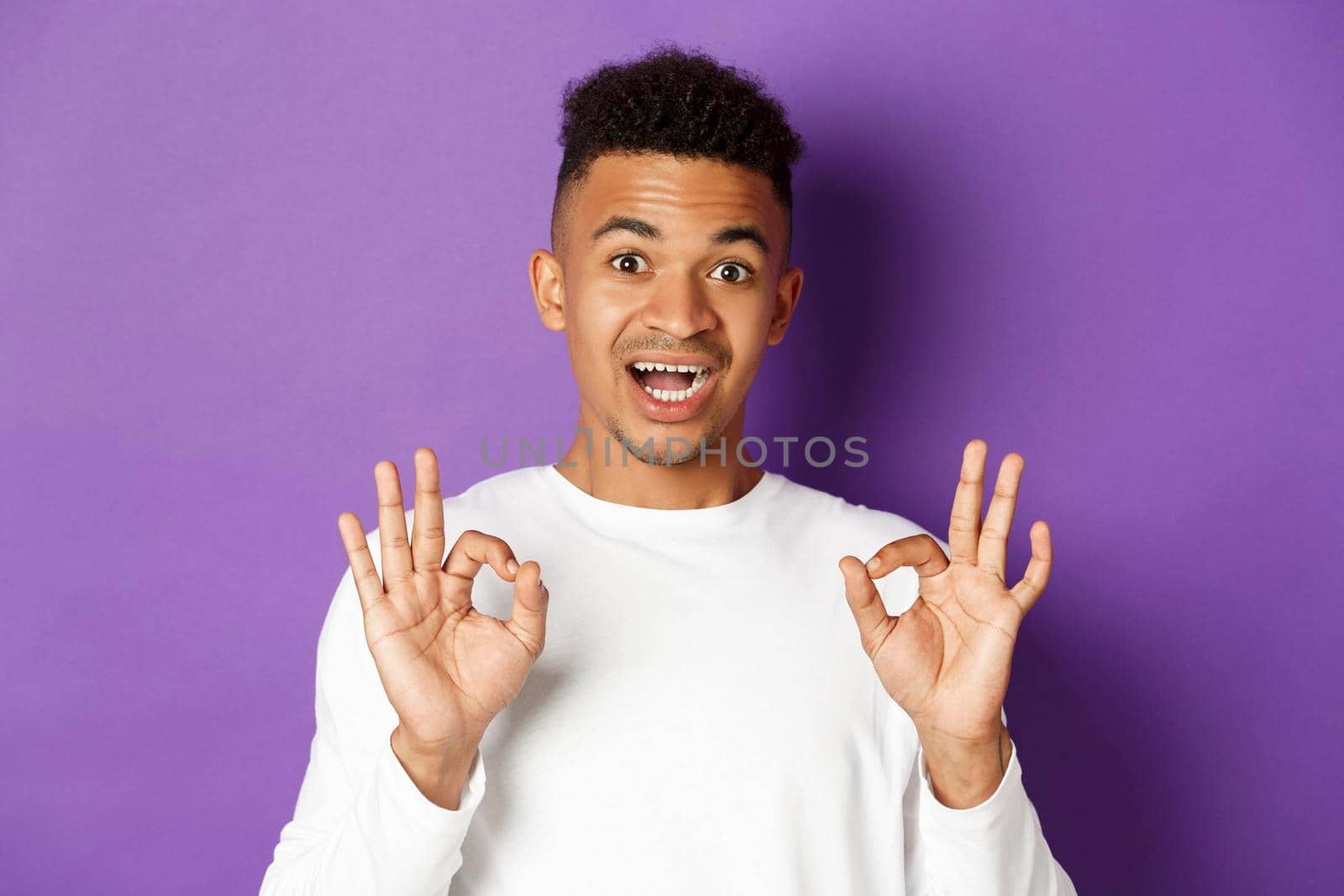 Close-up of handsome young african-american man, looking amazed and showing okay signs, recommend something good, standing over purple background by Benzoix