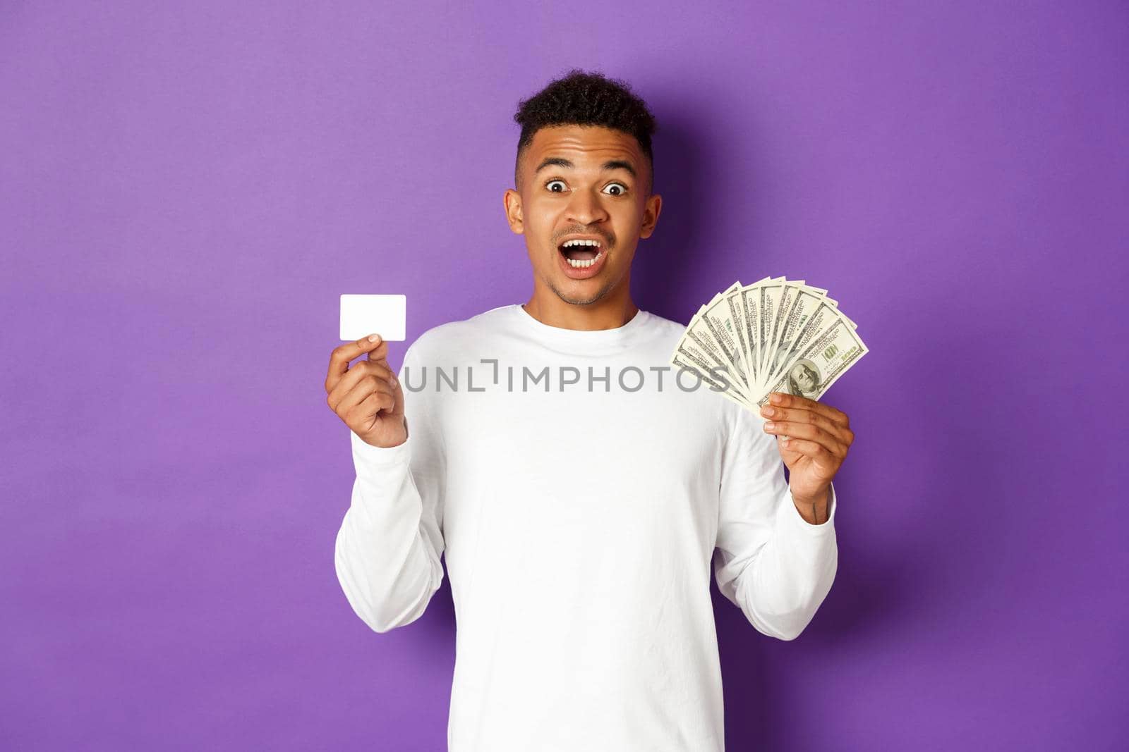 Image of handsome african american guy, looking pleased and smiling, showing credit card and money, standing over purple background.