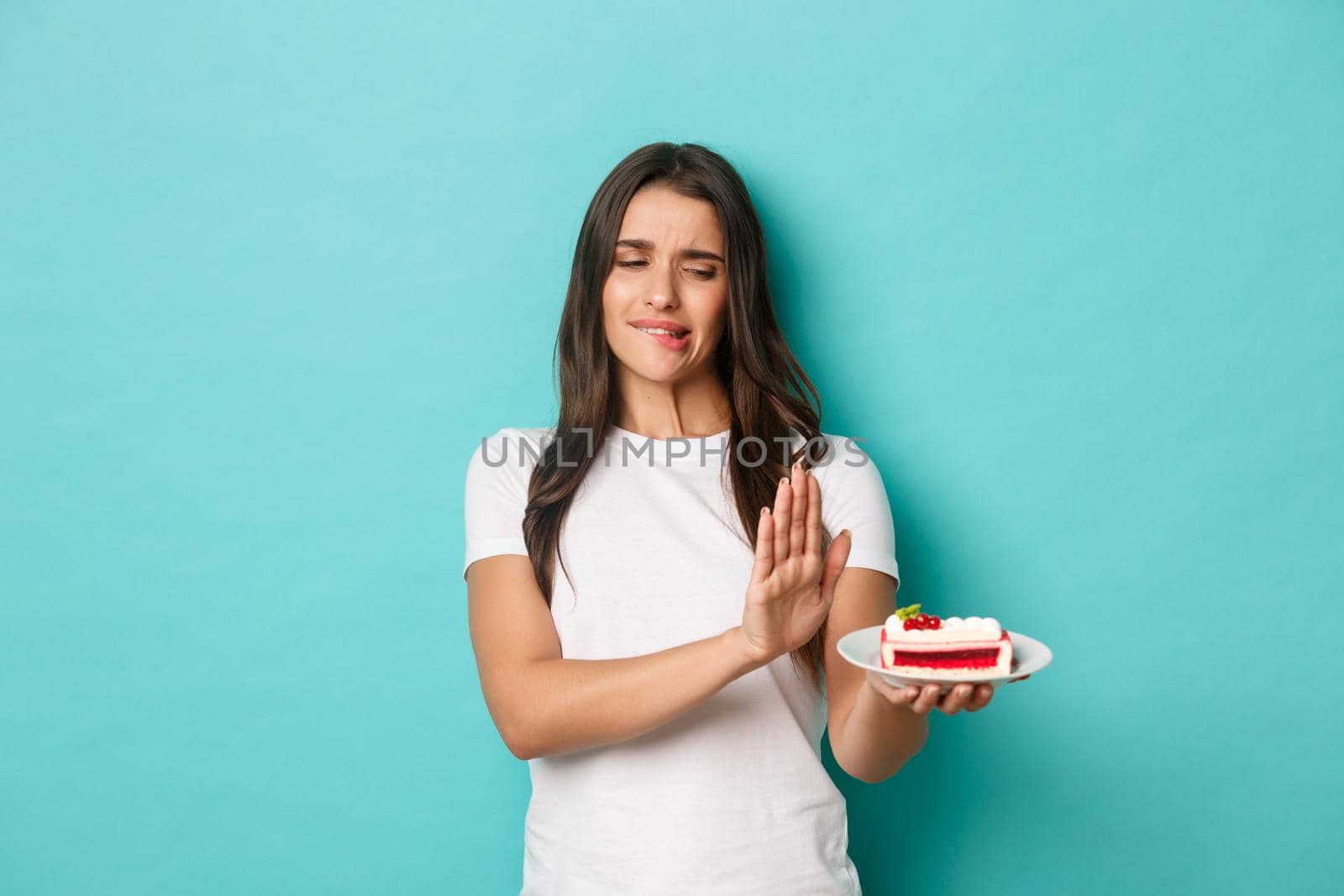 Image of cute girl on diet, tempted to eat cake but rejecting it, standing over blue background.