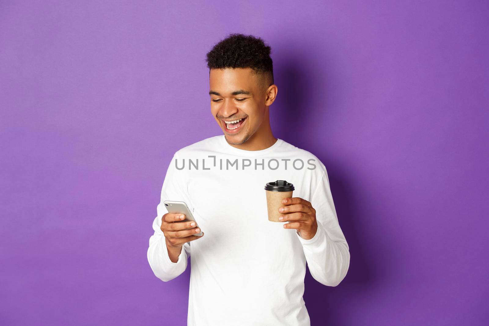 Happy african-american man drinking coffee, laughing and looking at smartphone, standing over purple background by Benzoix
