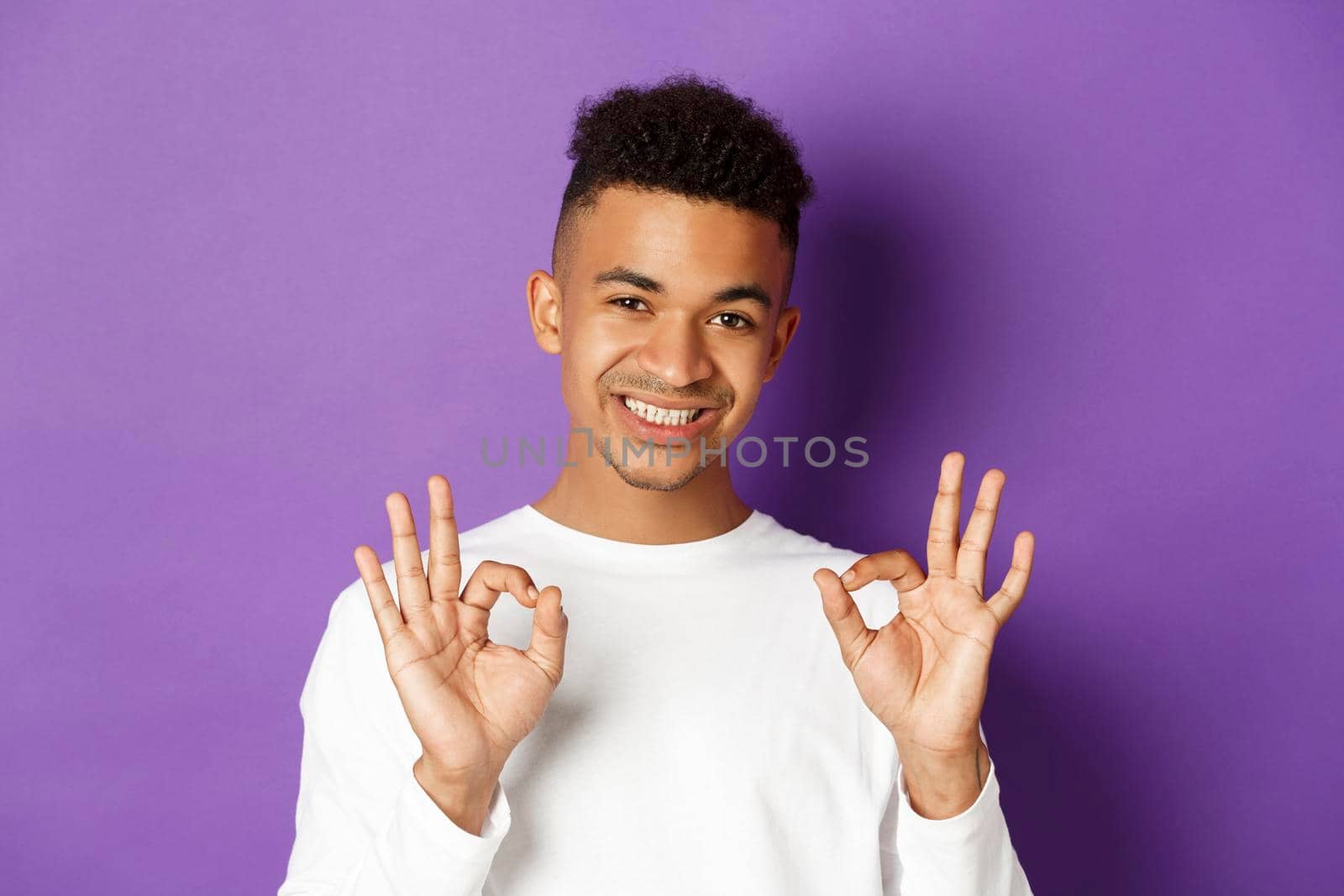 Close-up of young african-american guy looking satisfied, smiling and showing okay signs, approve and like something good, standing over purple background.