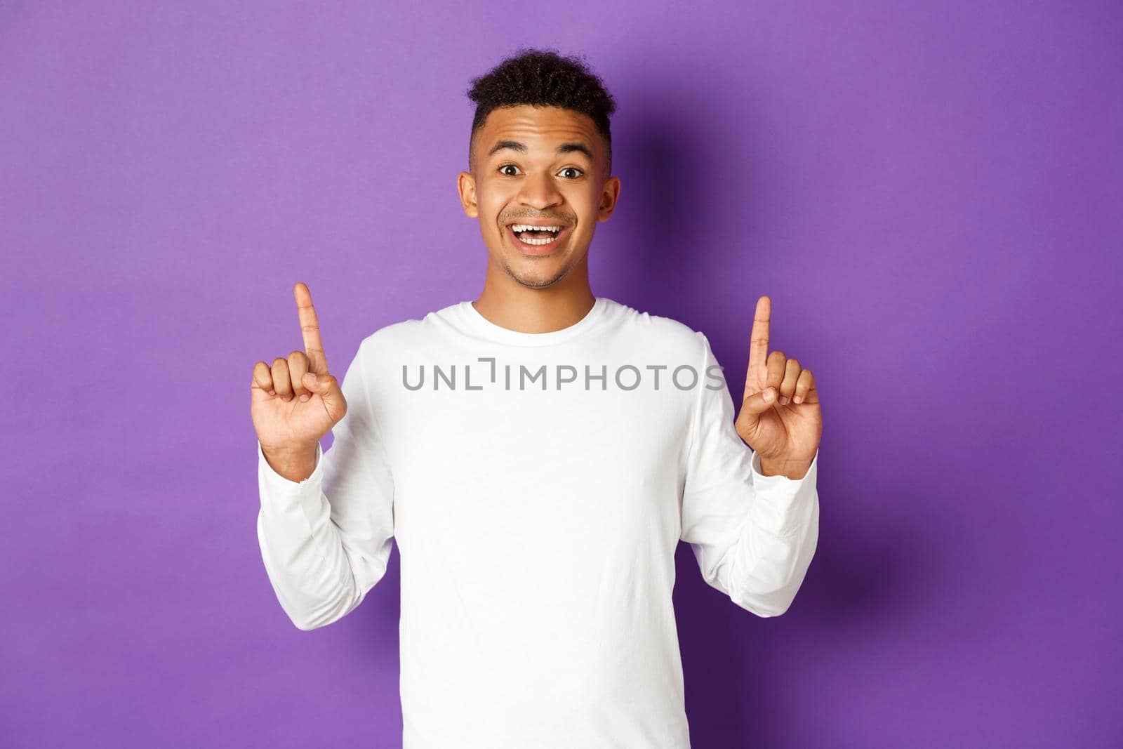 Cheerful african-american guy in white sweatshirt, pointing fingers up and smiling, making an announcement, showing your logo on copy space, standing over purple background.