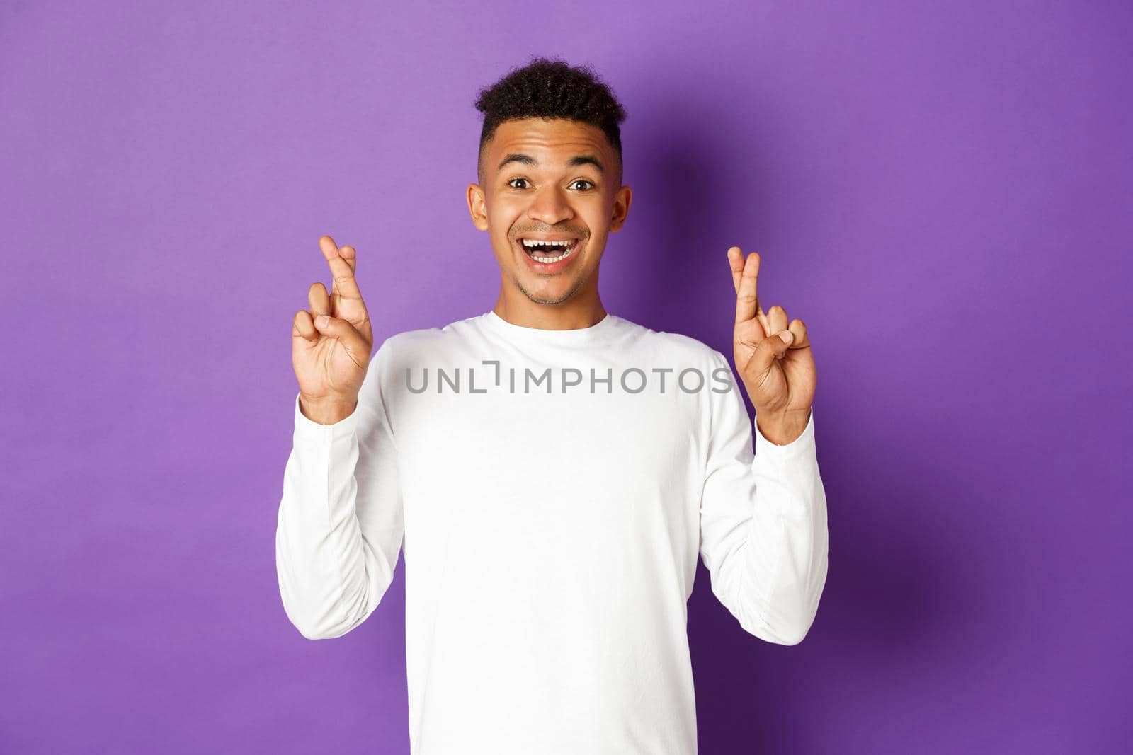 Image of handsome african-american young guy in white sweatshirt, looking hopeful and smiling with crossed fingers, making wish or waiting for good news, standing over purple background.