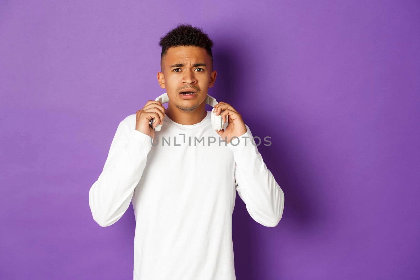 Image of modern african-american man, take-off headphones and looking confused, standing over purple background by Benzoix