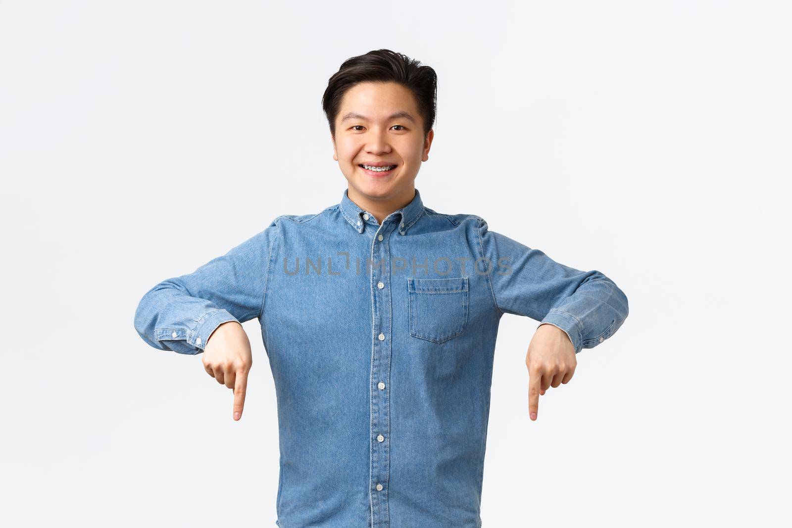 Smiling confident asian man satisfied with his stomatology clinic, recommend doctor, showing braces proud, pointing fingers down satisfied, standing white background. Copy space