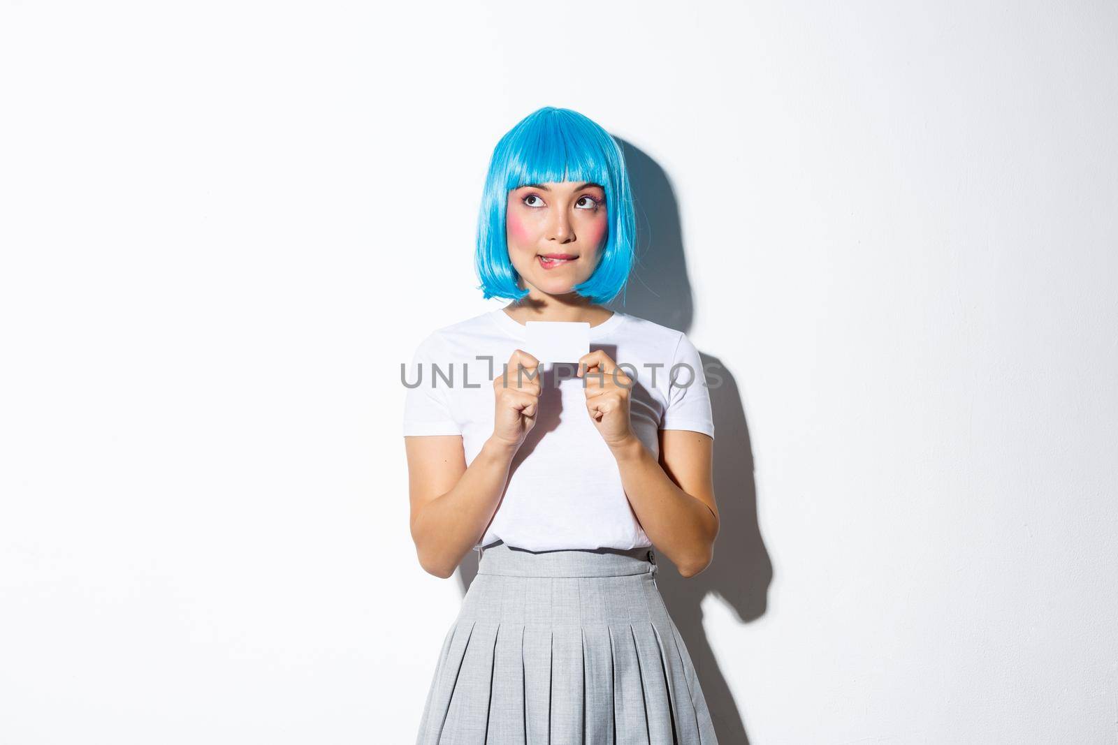 Image of thoughtful cute asian girl in blue wig making choice, looking upper left corner and showing credit card by Benzoix