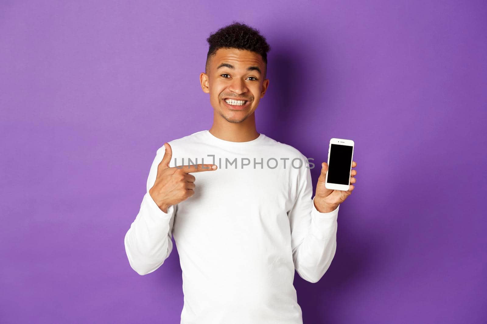 Portrait of handsome african-american man, smiling and pointing finger at smartphone screen, showing application, standing over purple background.
