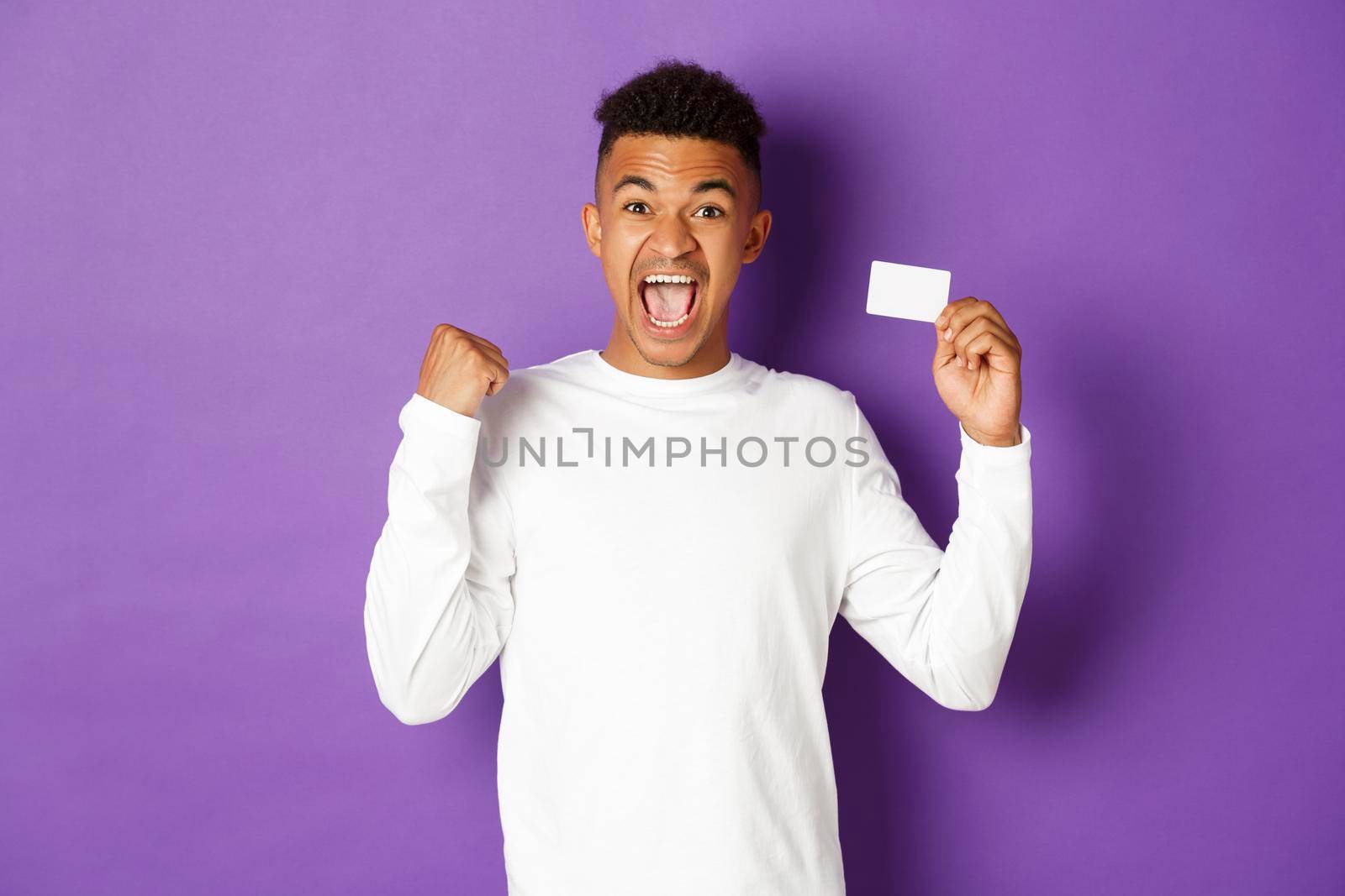 Portrait of handsome african-american man, looking excited and cheerful, showing credit card and making fist pump gesture, standing over purple background by Benzoix